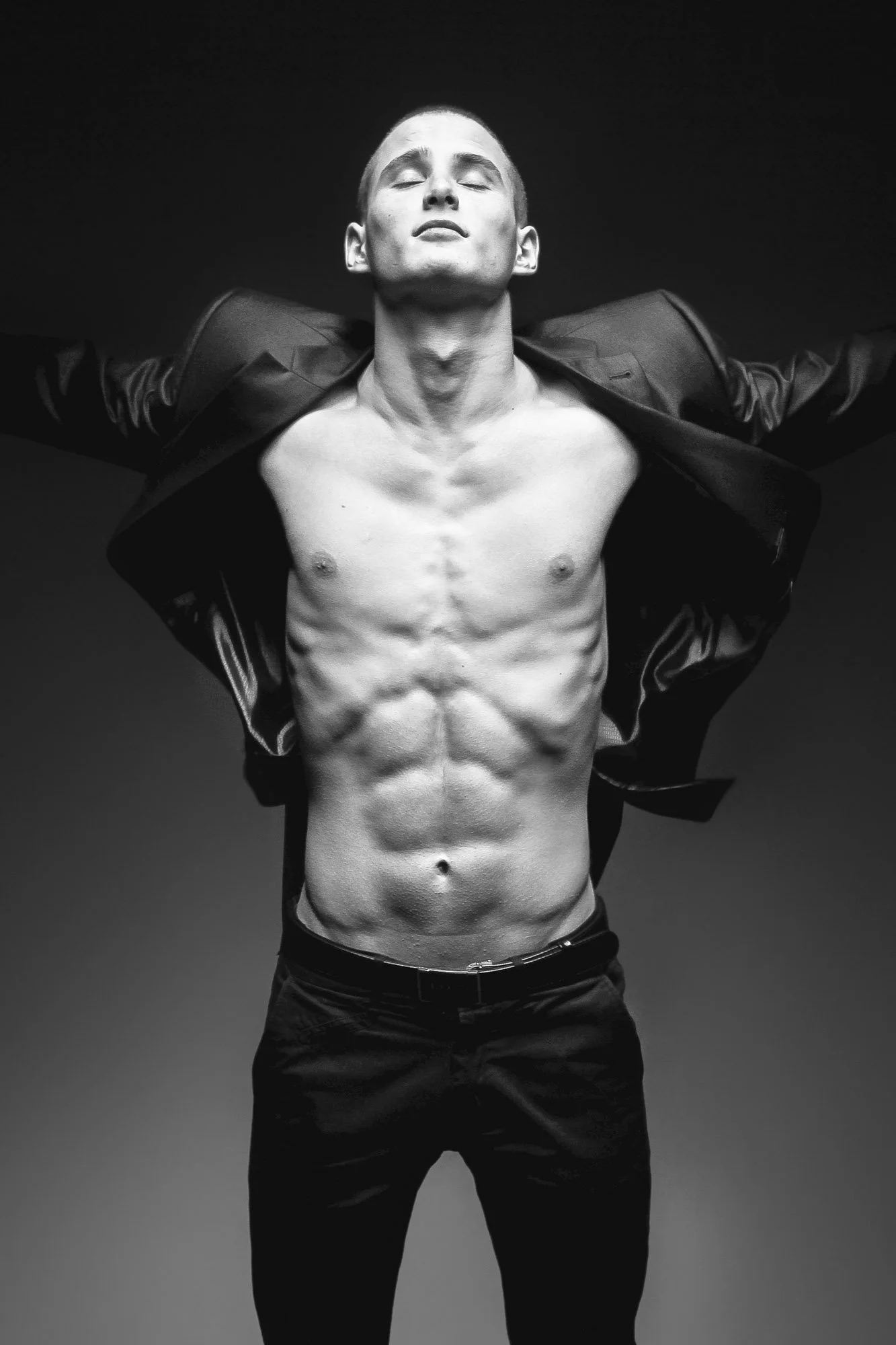 Studio male portrait, man. Artistic male portrait photography Zurich. Black and white photo of a shirtless young man with his eyes closed, arms extended outwards, wearing an open jacket and black pants.