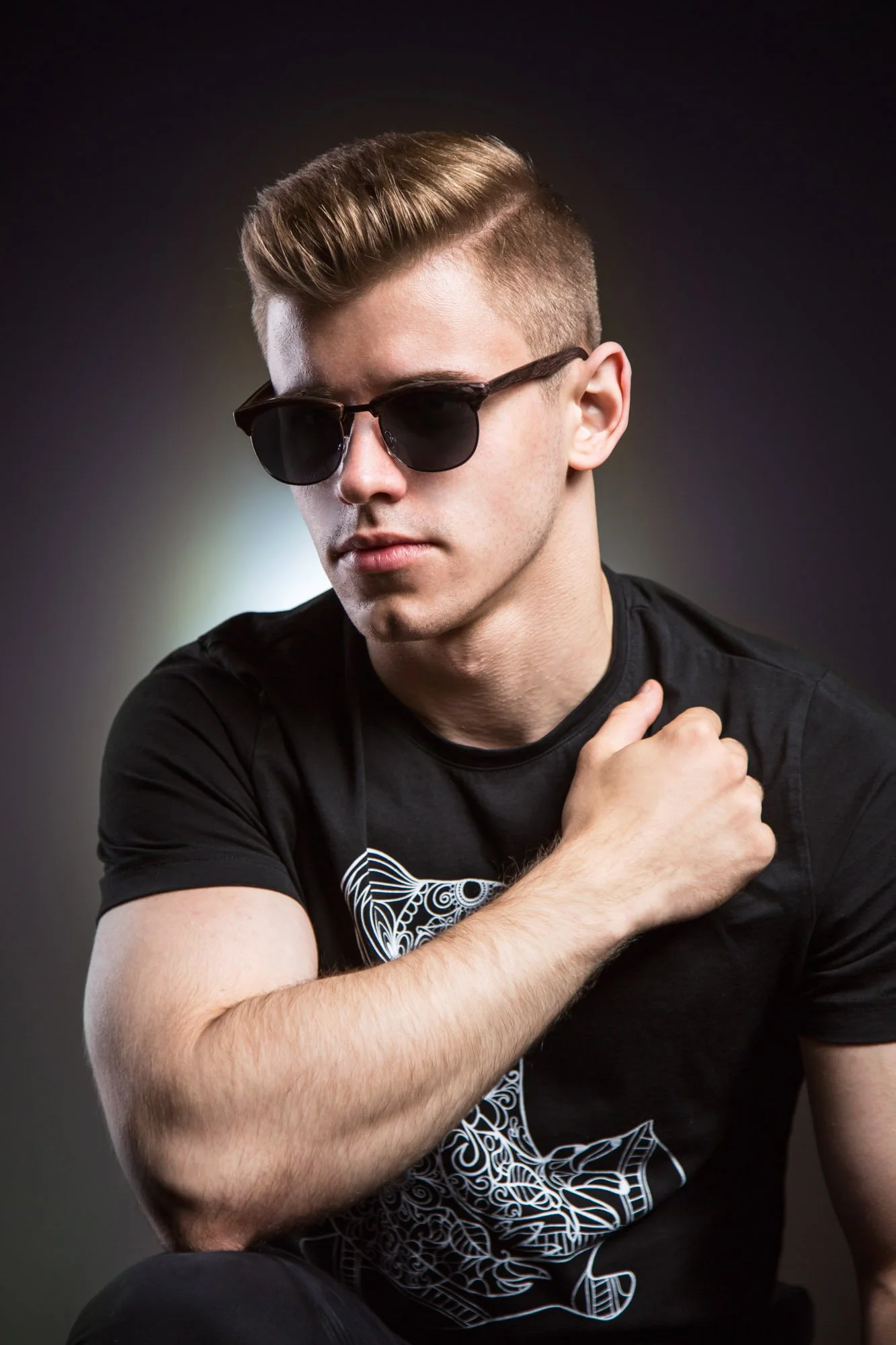 Portrait of a young man wearing black sunglasses, a black t-shirt with a white graphic design, and touching his shoulder with one hand, set against a dark gradient background.