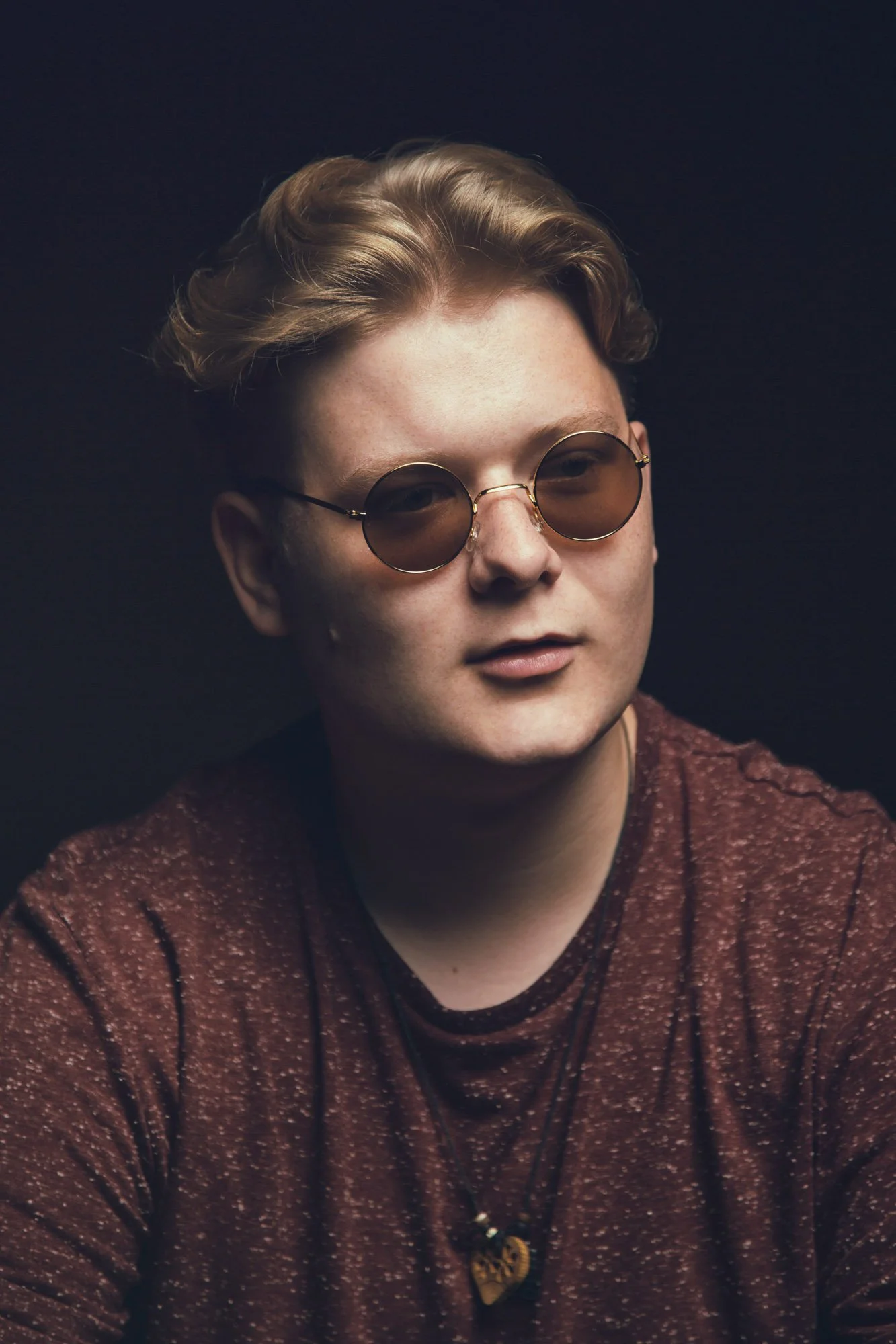 A young man with wavy blond hair wearing round sunglasses and a brown speckled shirt, with a dark background.