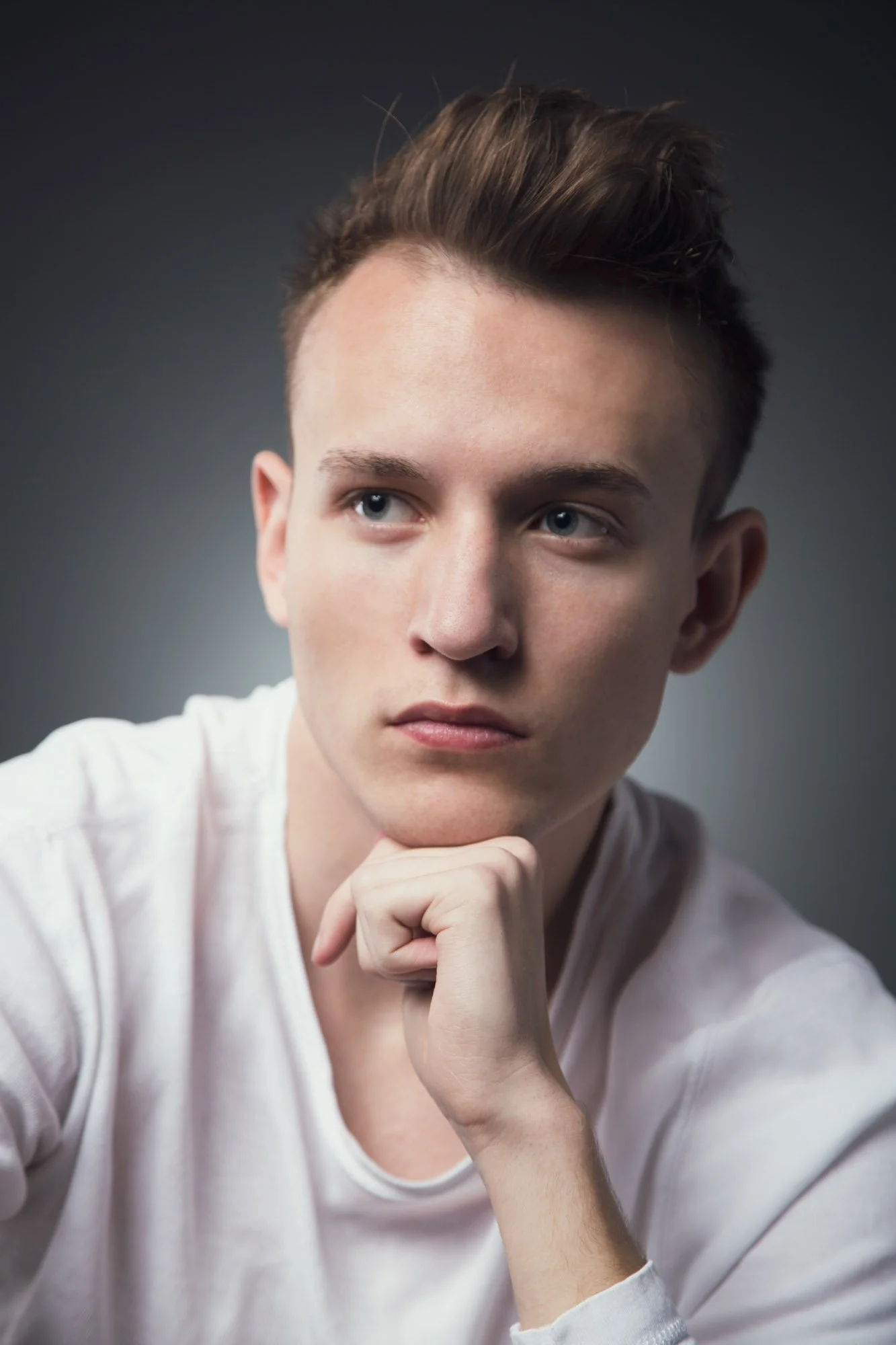 Portrait, Fitness & Commercial Photographer Zurich. Close-up of a young man with styled brown hair, fair skin, and blue eyes, resting his chin on his hand, looking to the side against a dark gray background.