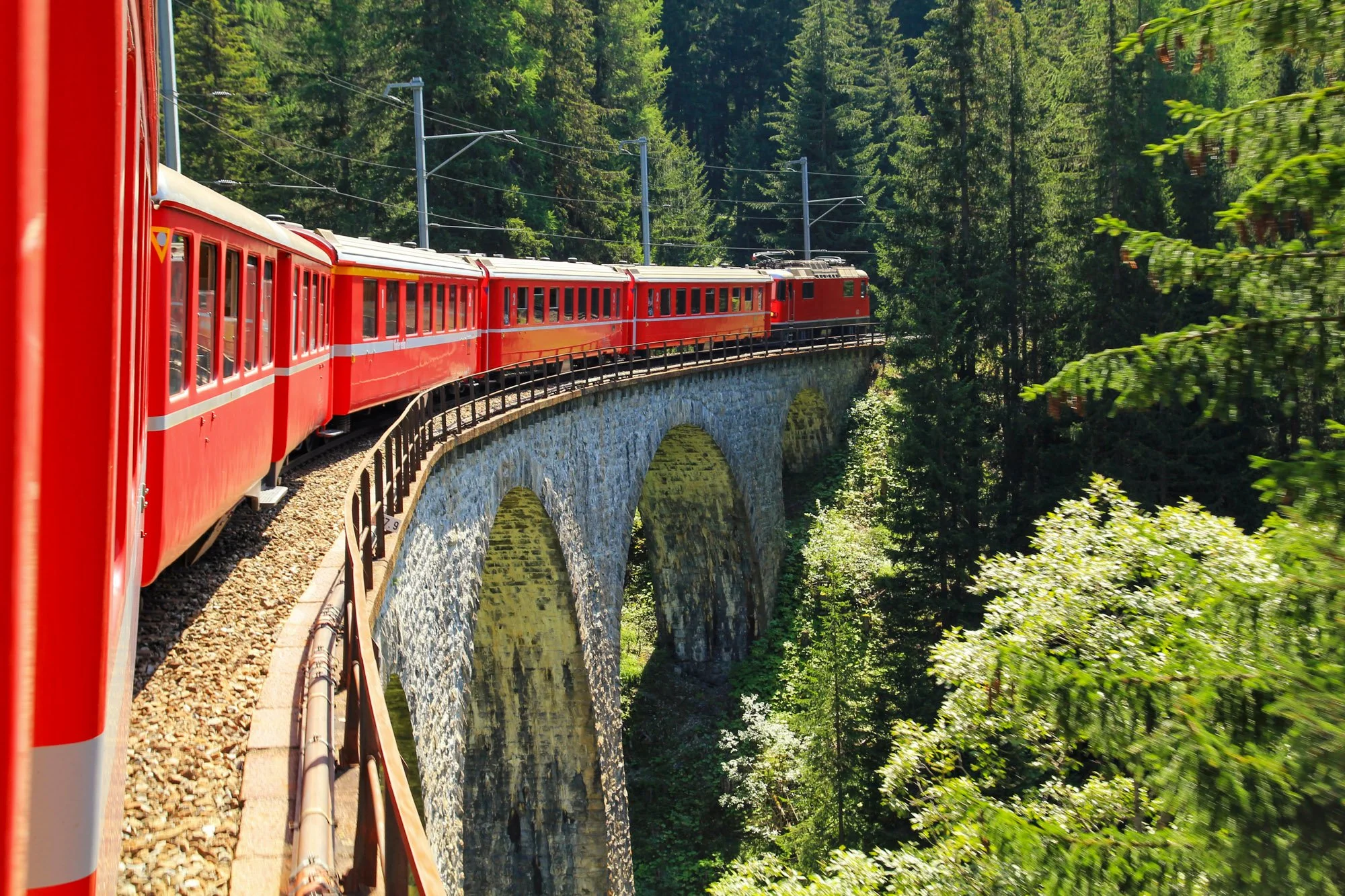 Fine art print photography for modern living room wall decor in Zürich. A red train traveling over a stone arch bridge surrounded by lush green forest.
