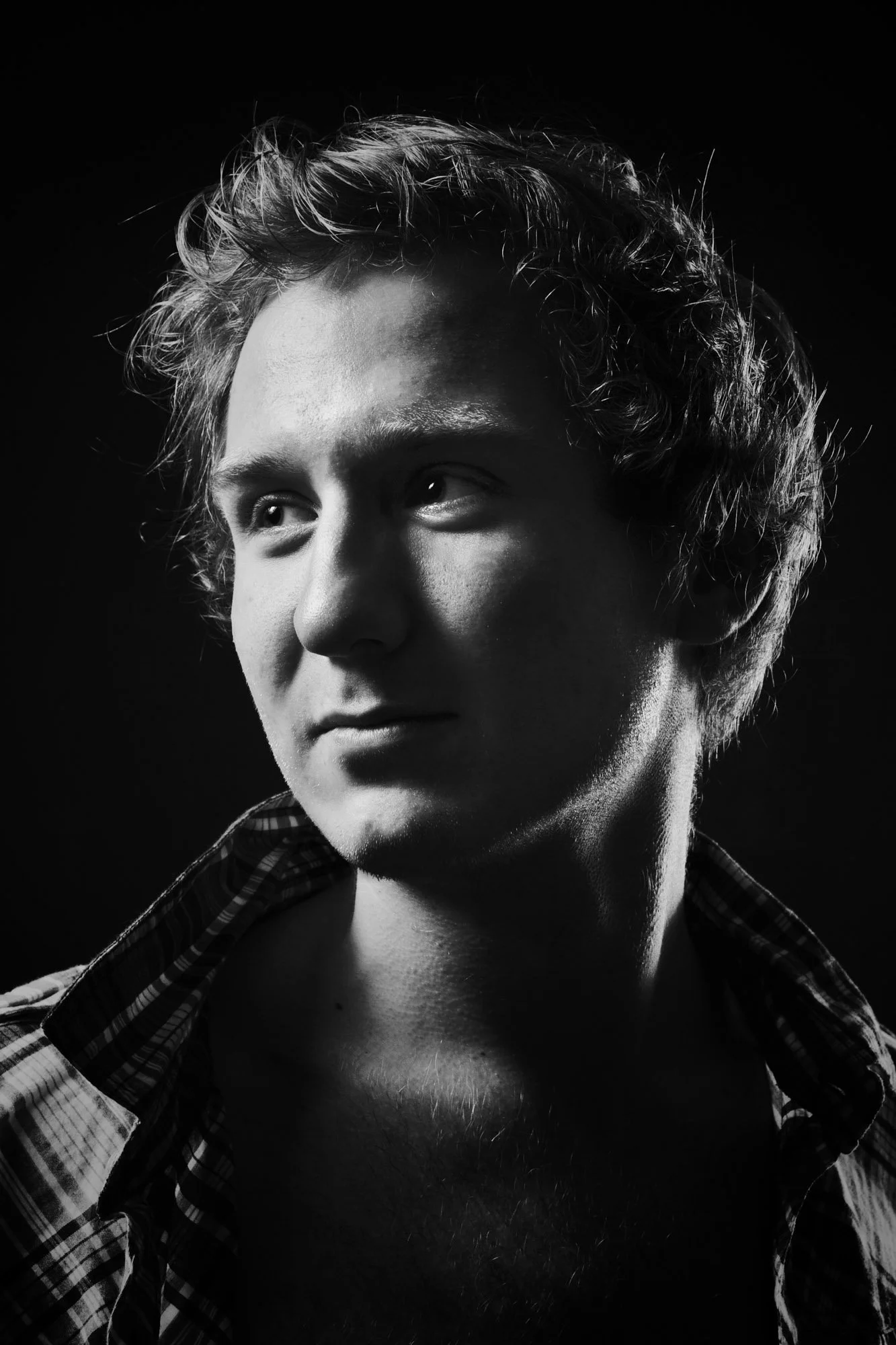Black and white portrait of a young man with curly hair, wearing an unbuttoned plaid shirt, looking to the side.
