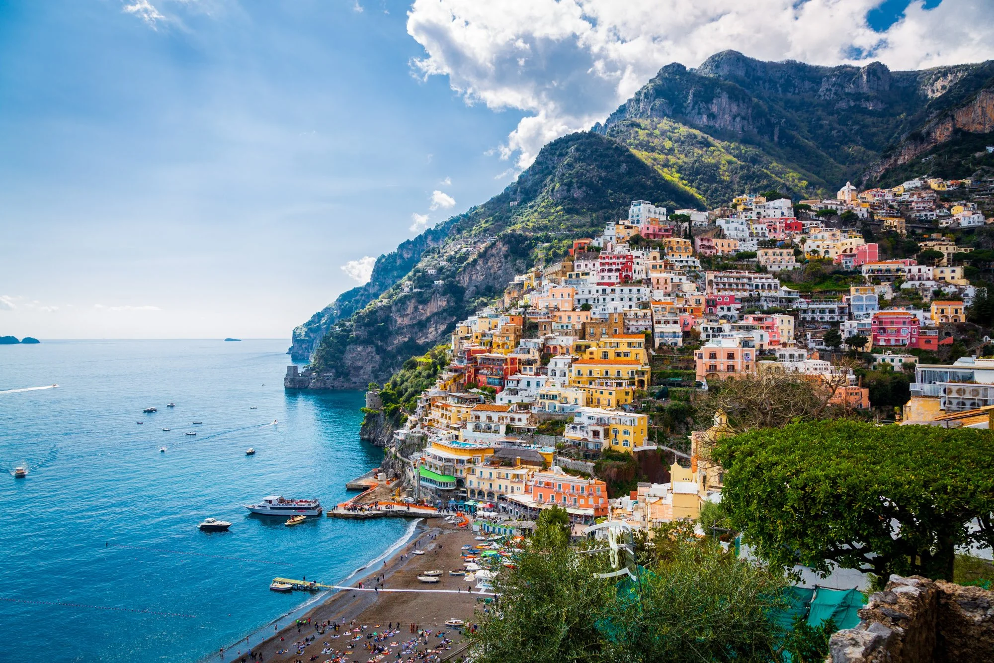 Colorful buildings on a hillside overlooking the ocean, with boats in the water and a beach below, under a partly cloudy sky.