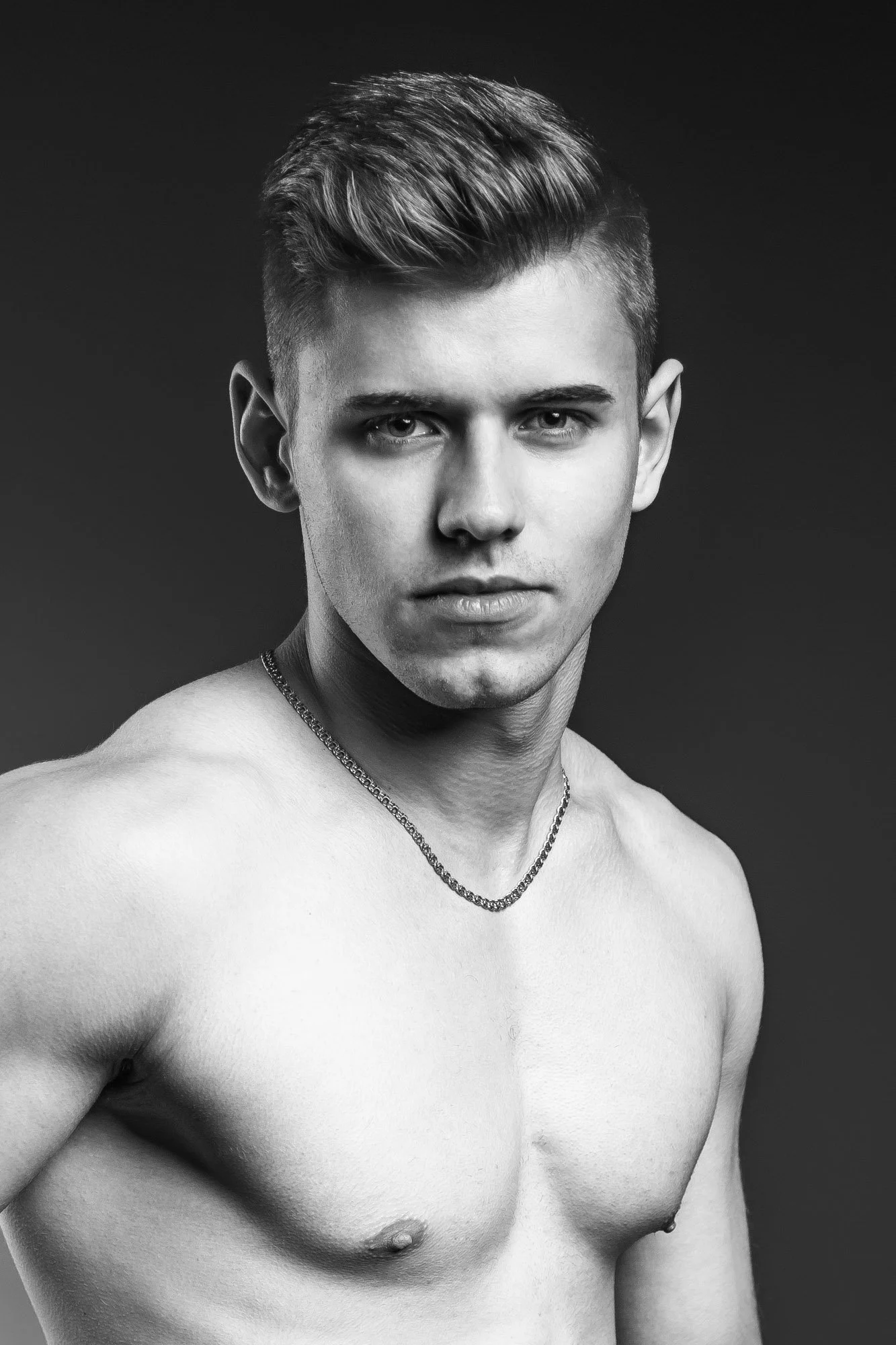 Black and white portrait of a young man with a serious expression, short styled hair, shirtless showing chest and shoulder, wearing a chain necklace, against a plain dark background.