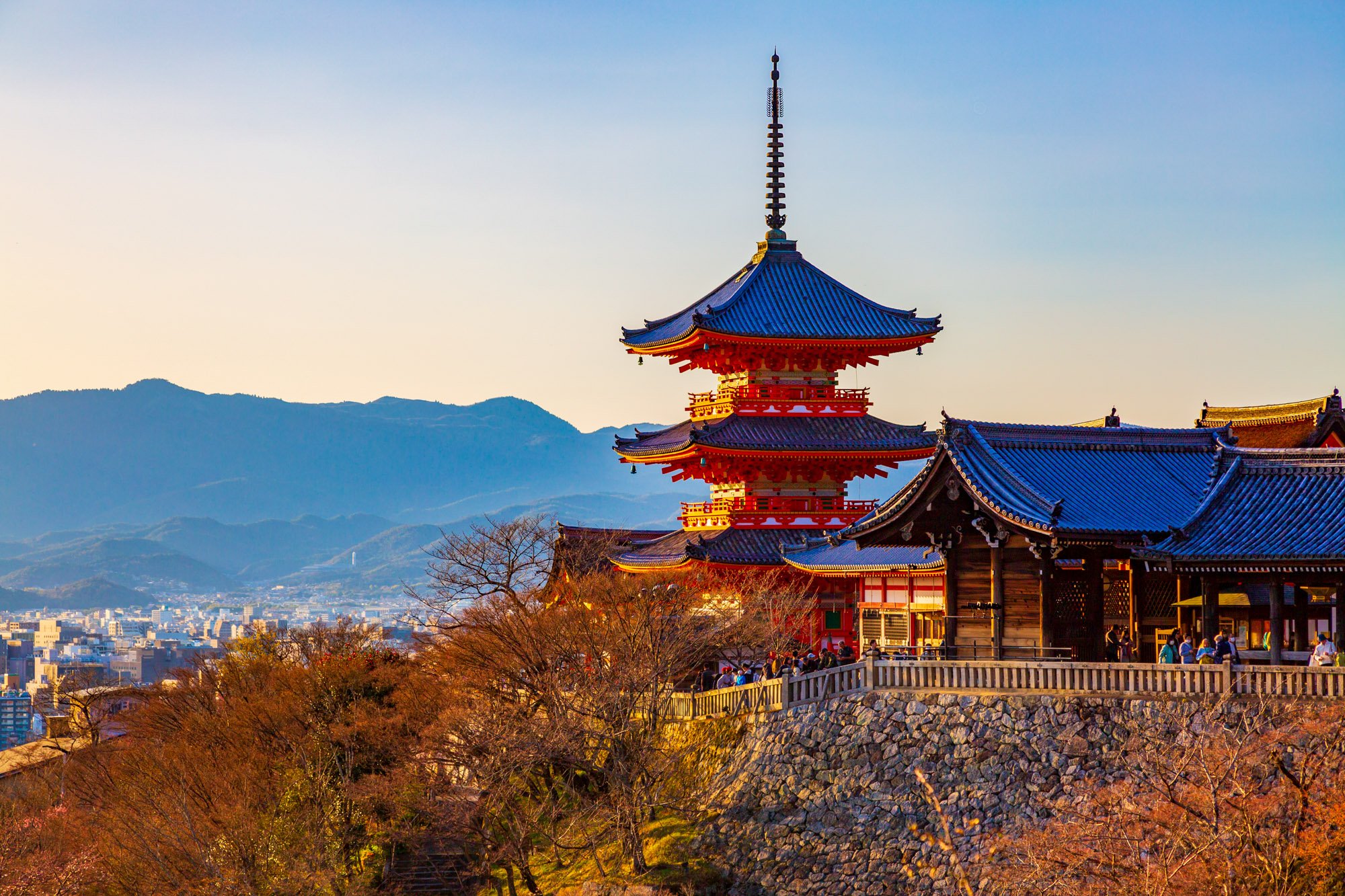 Traditional Japanese pagoda with blue roof, red structure, and ornate details, situated on a hilltop with a scenic view of mountains in the background, surrounded by trees under a clear sky.