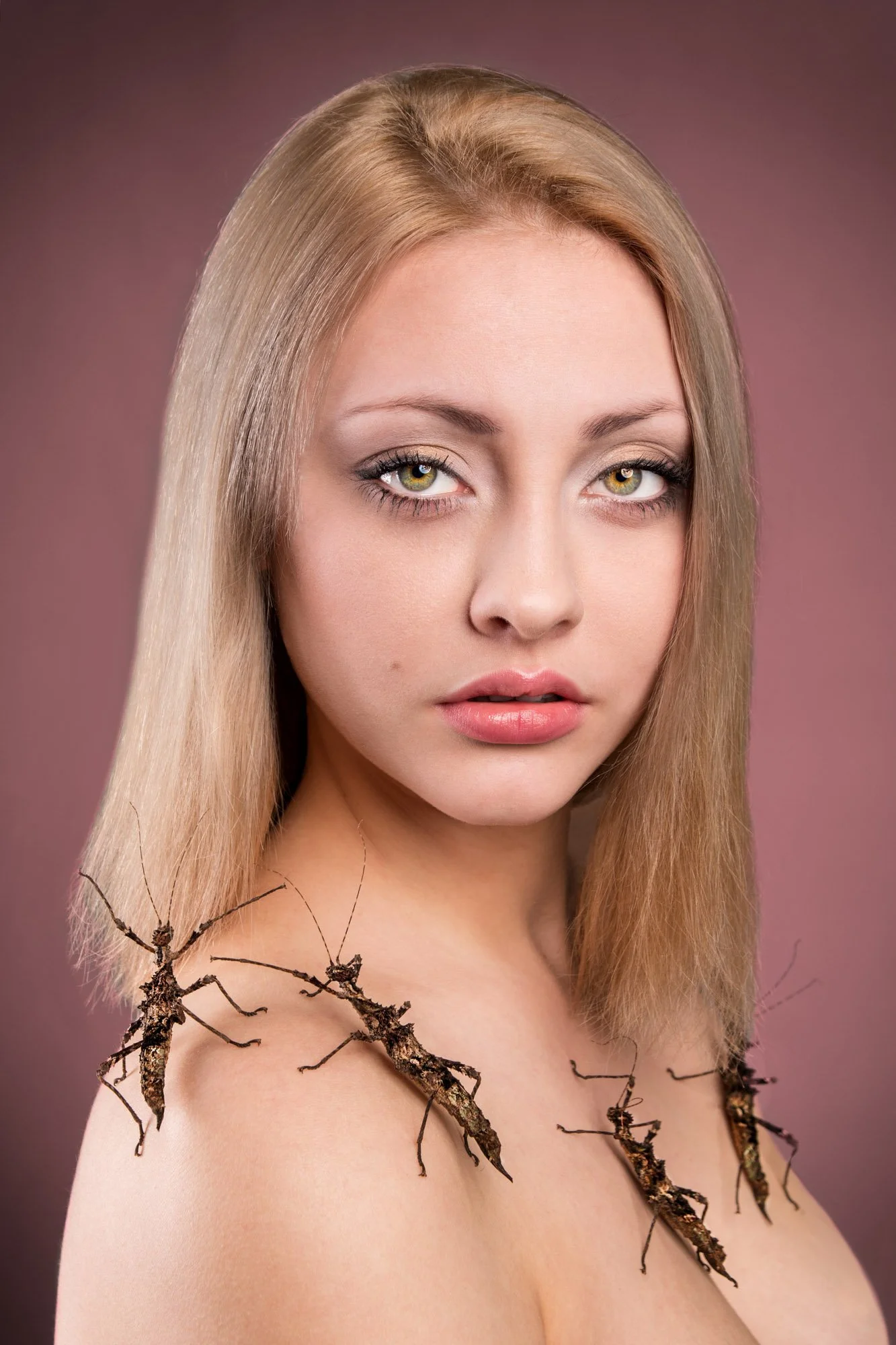 Portrait of a young woman with blonde hair, green eyes, and natural makeup, wearing a necklace made of intertwined twigs and branches against a soft pink background.