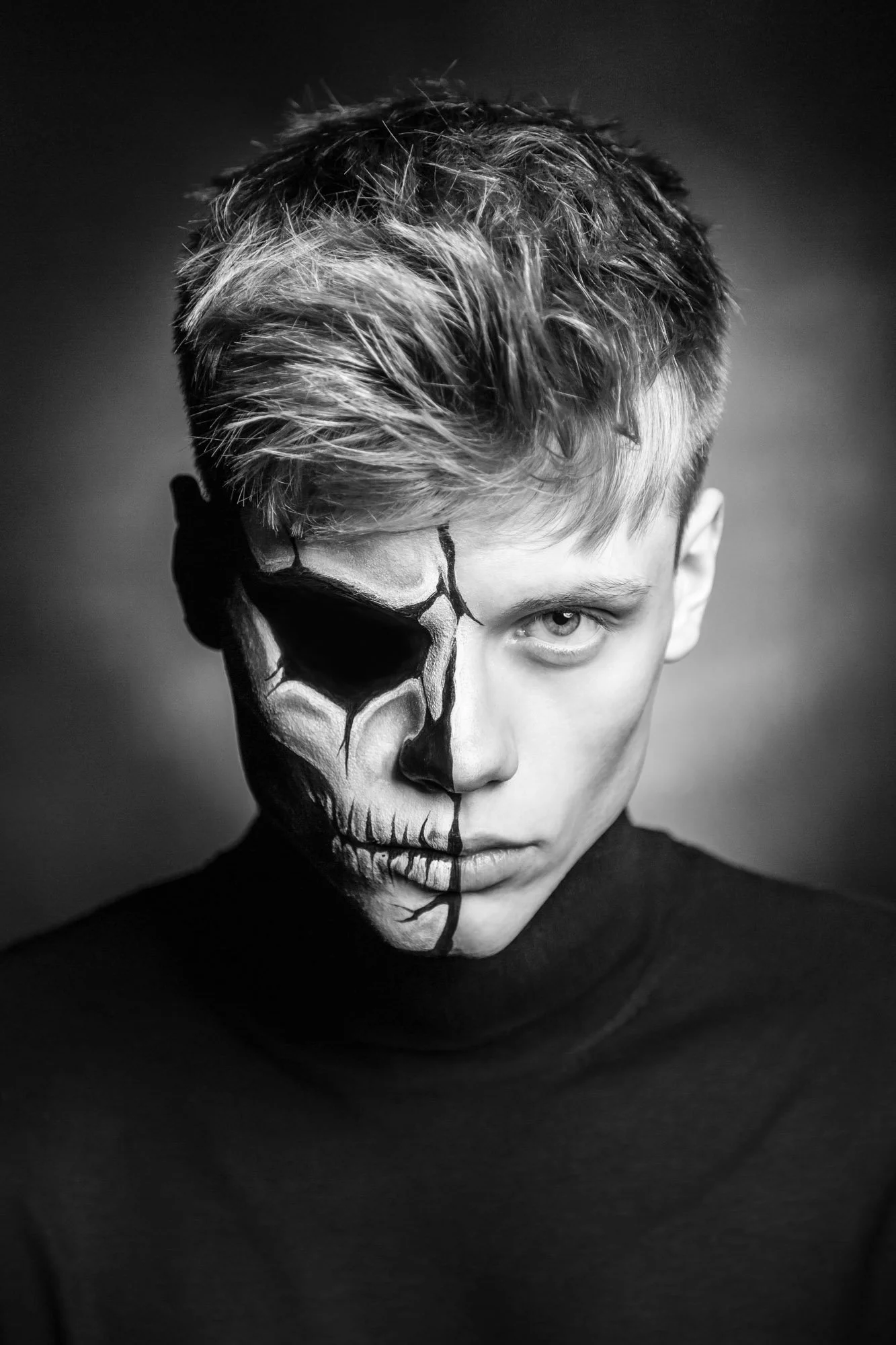 Artistic male portrait photography Zurich. Black and white portrait of a young man with half his face painted as a skull, with intense expression and styled hair, wearing a dark turtleneck.