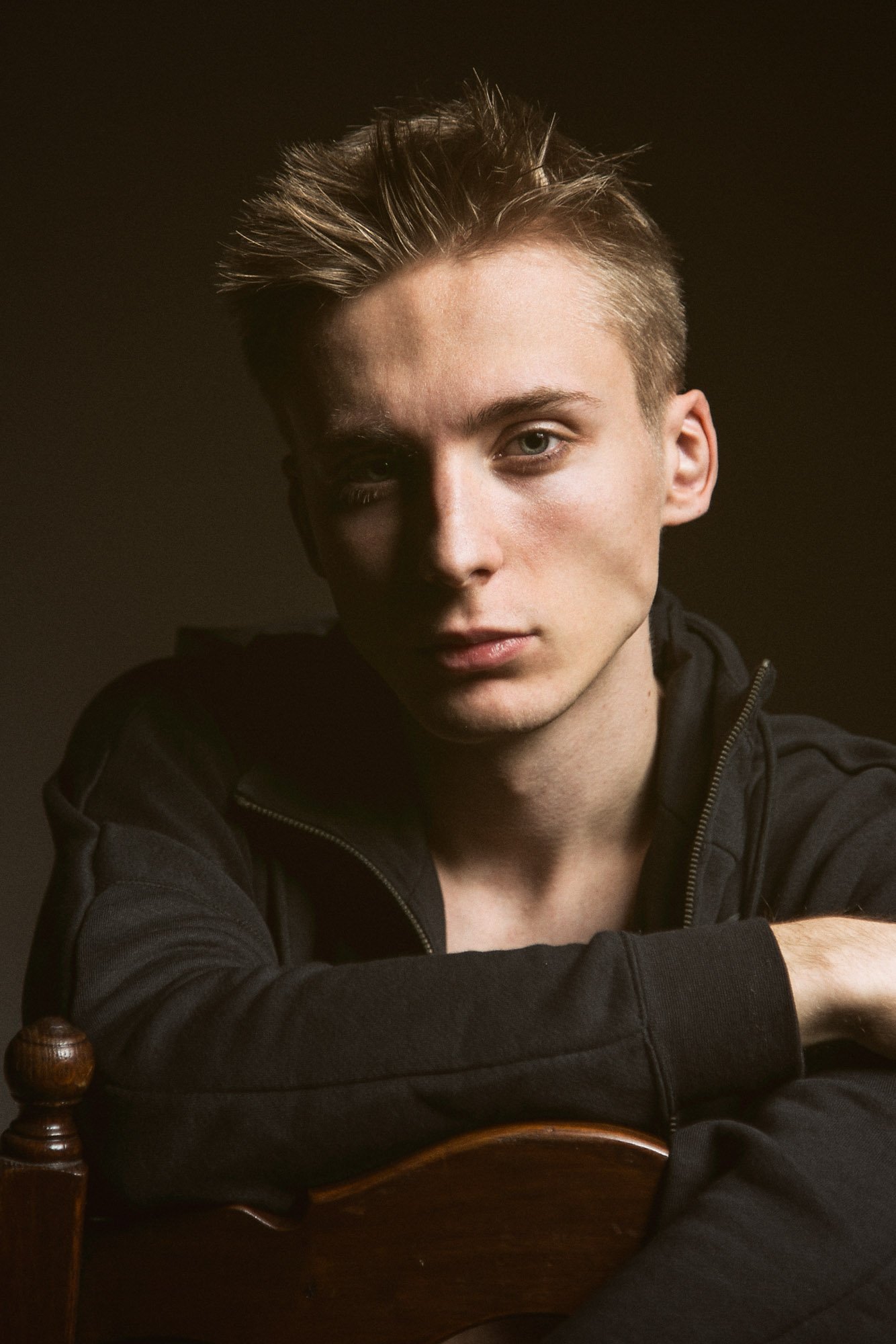 Portrait, Fitness & Commercial Photographer Zurich. A young man with light skin and styled hair looking at the camera, sitting with his arms crossed on a wooden chair, wearing a black zip-up jacket, against a dark background.