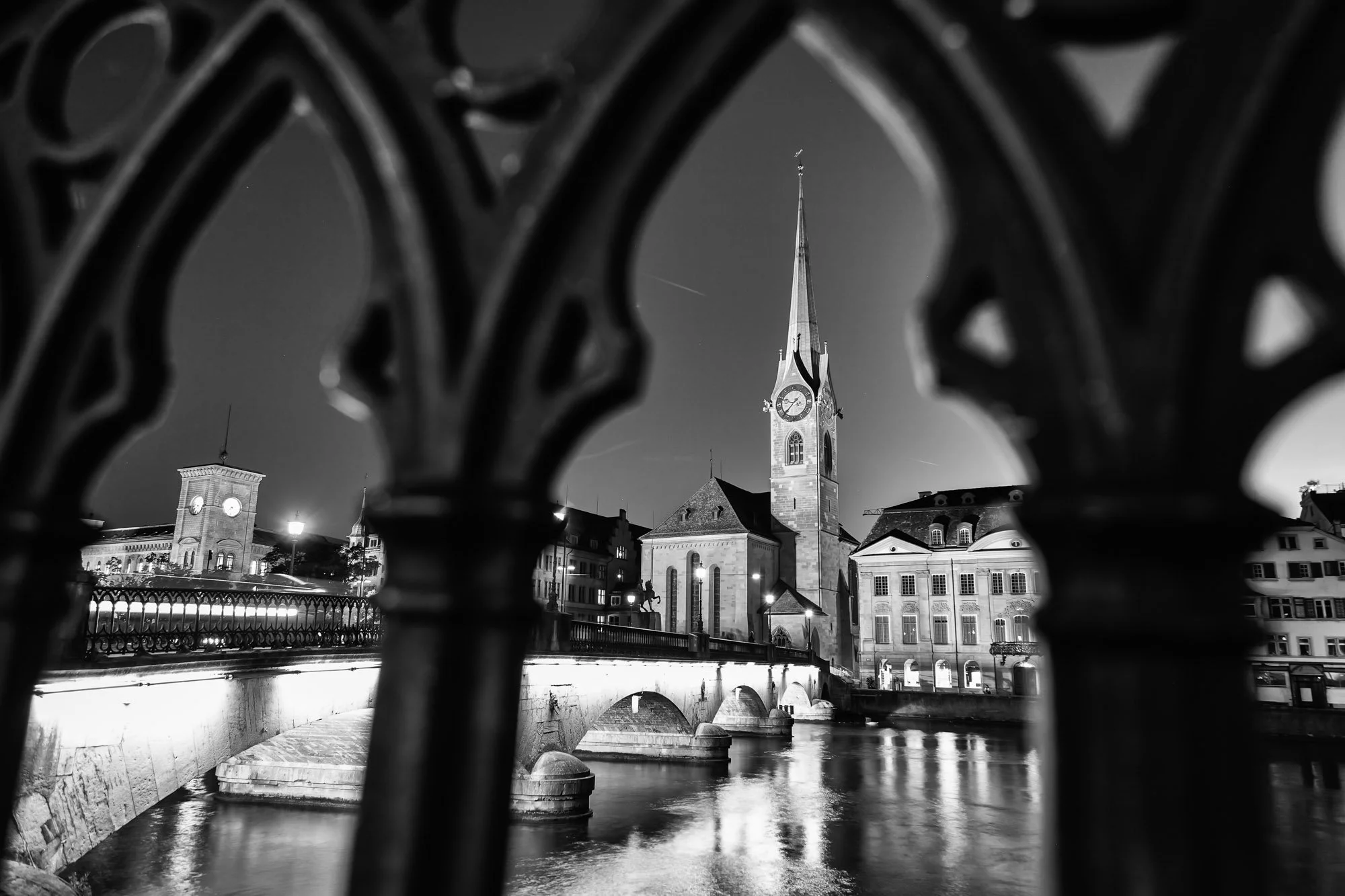 Fine art print photography for modern living room wall decor in Zürich.. Nighttime view of a church with a tall steeple and clock, seen through decorative iron railings over a bridge crossing a river.