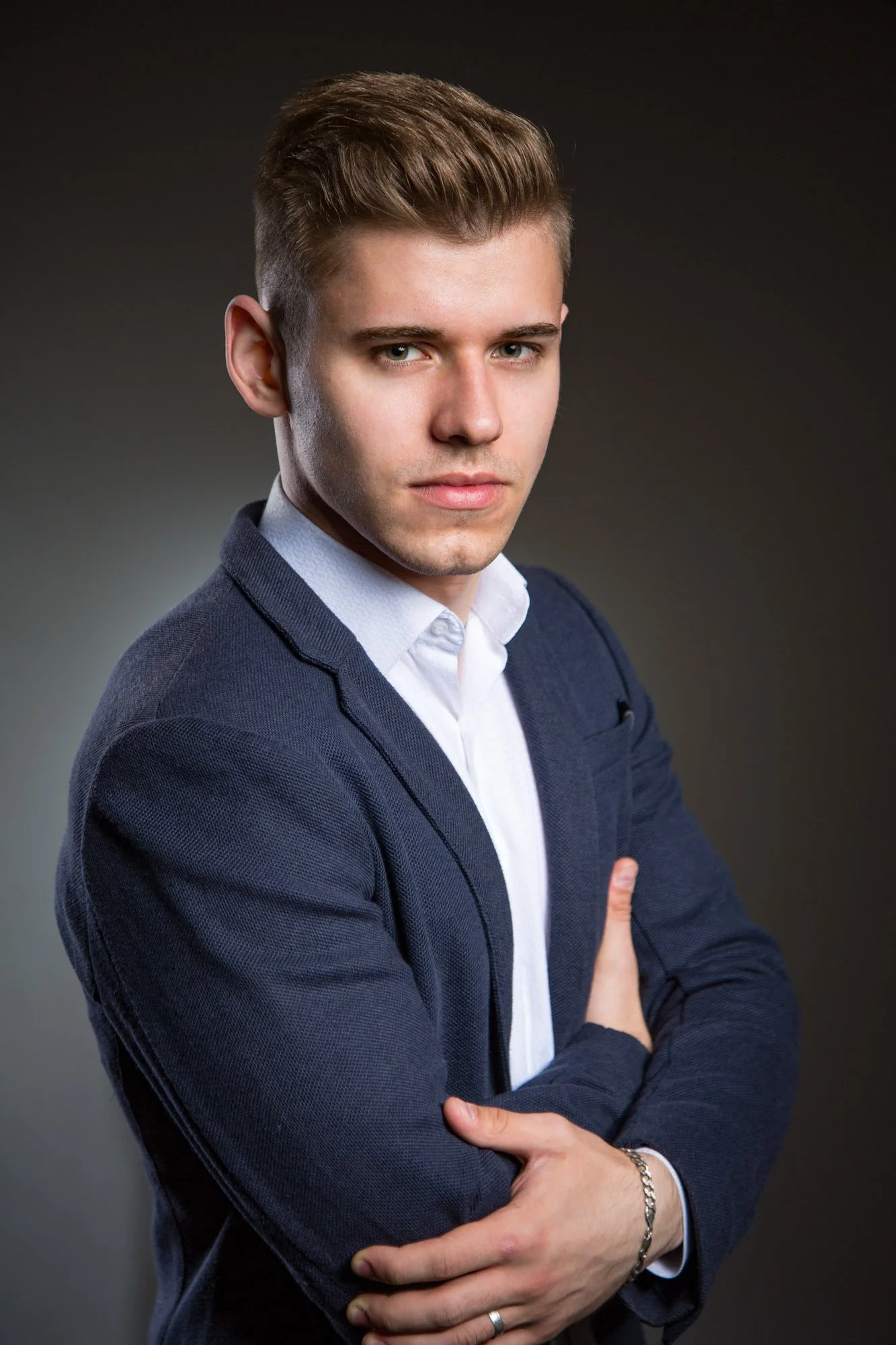 Portrait, Fitness & Commercial Photographer Zurich. Young man with short brown hair, wearing a navy blazer and white shirt, crossing his arms in front of a dark background.