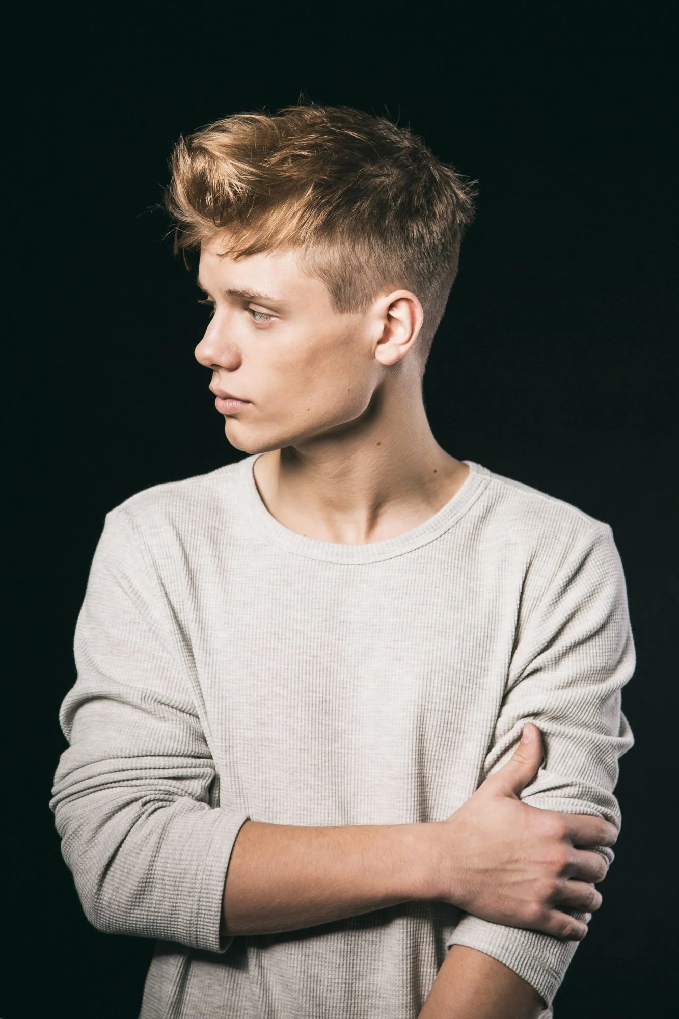 A young man with short, styled hair, wearing a light-colored sweatshirt, standing with crossed arms against a black background.