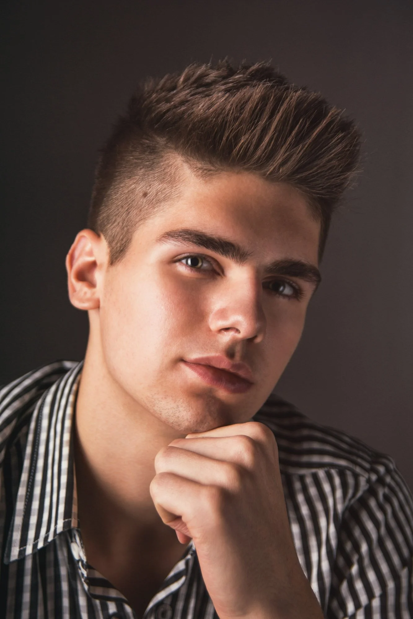 Portrait, Fitness & Commercial Photographer Zurich. Close-up portrait of a young man with styled brown hair, wearing a striped shirt, resting his chin on his hand, against a dark background.