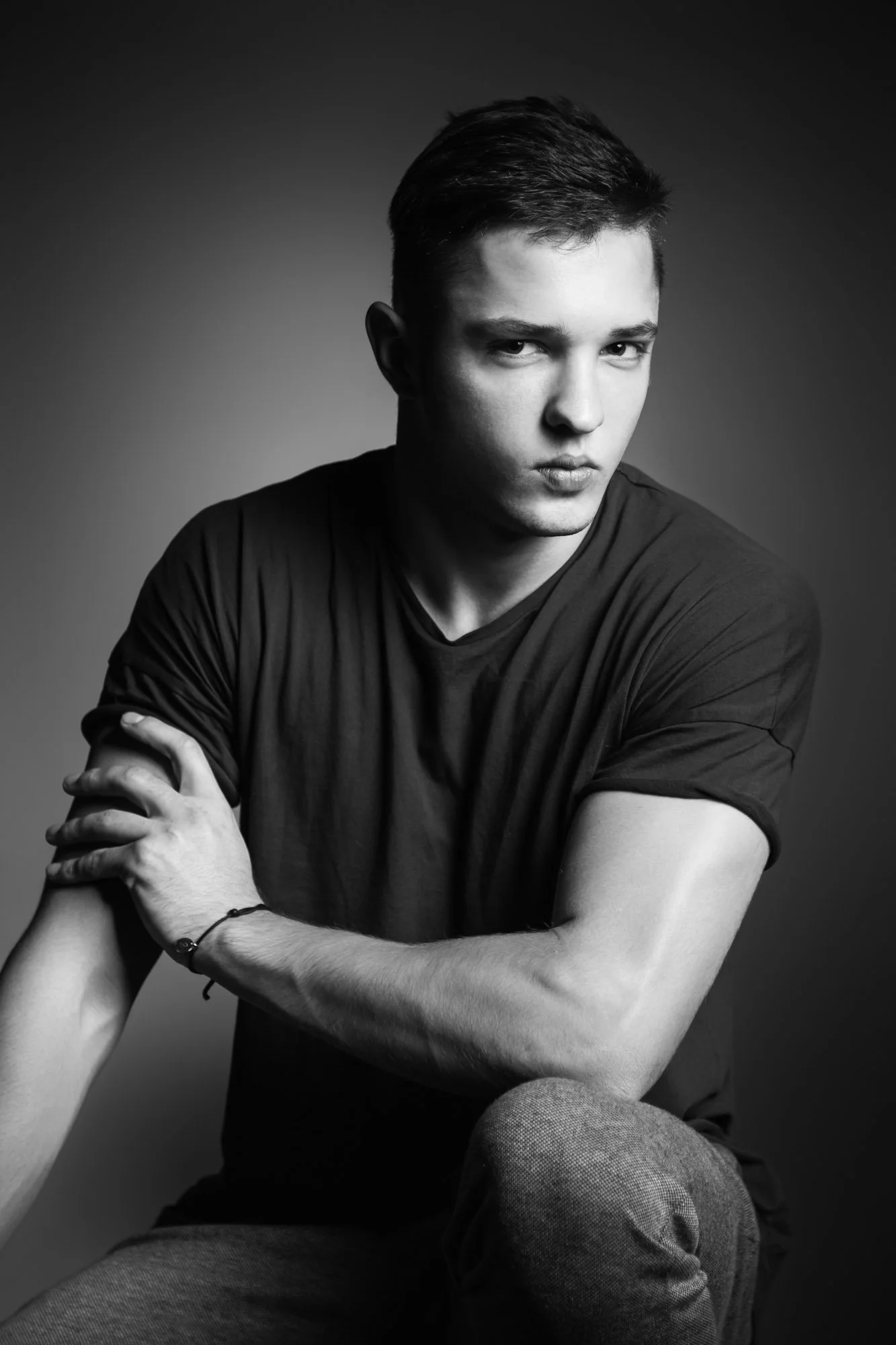 Black and white portrait of a young man with short hair, looking at the camera with a serious expression, sitting with arms crossed against a gradient background.