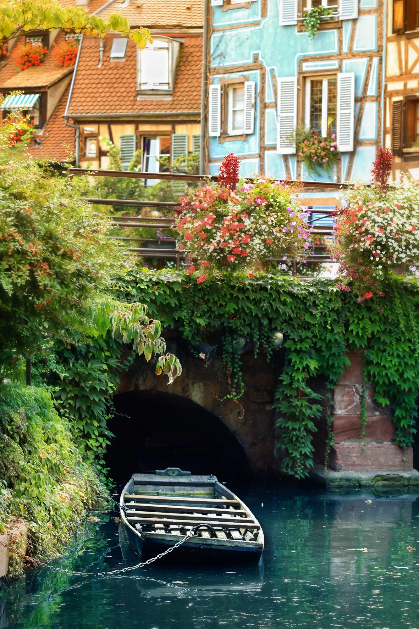A small boat is tied to the edge of a canal, with lush green plants and colorful flowers, leading to a tunnel under old buildings with flower boxes and shutters.