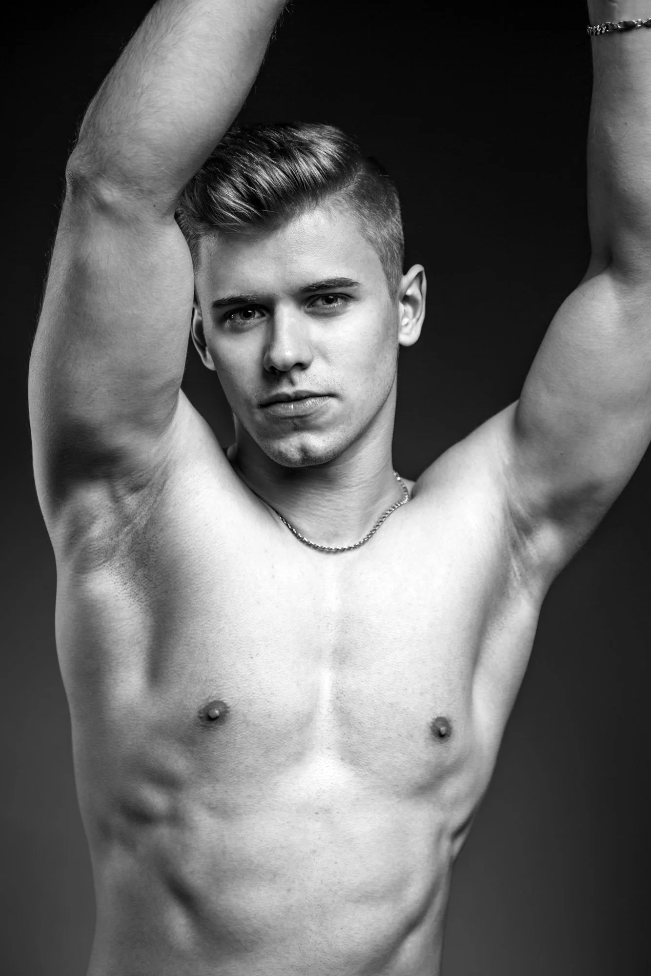 Black and white portrait of a young shirtless man with short hair, looking at the camera, in front of a dark background.