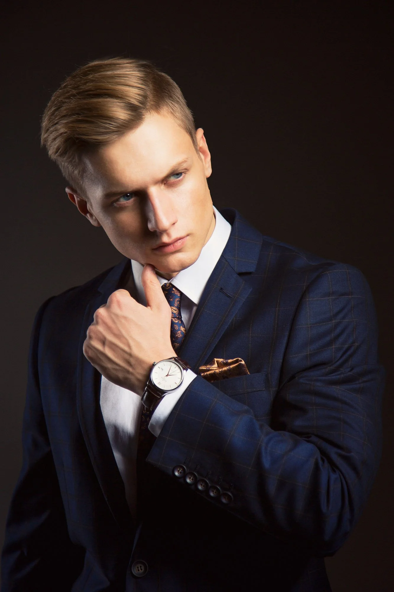 Portrait, FitnessA young man with blond hair styled to the side, wearing a dark blue checkered suit, white shirt, and patterned tie, looking thoughtfully to the side with his hand near his face. He is wearing a silver wristwatch with a leather strap.