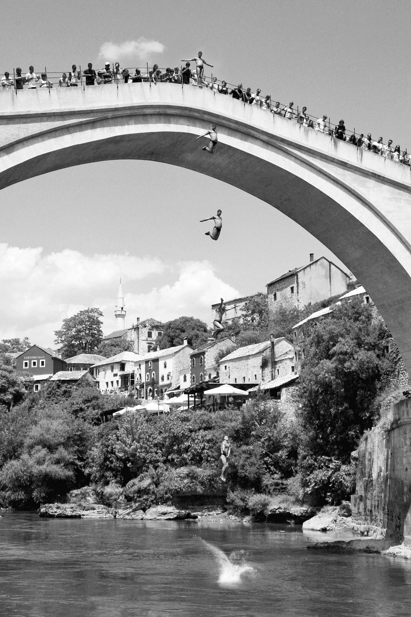 People jumping off a bridge into a river, with onlookers watching from the bridge and buildings on the hillside.