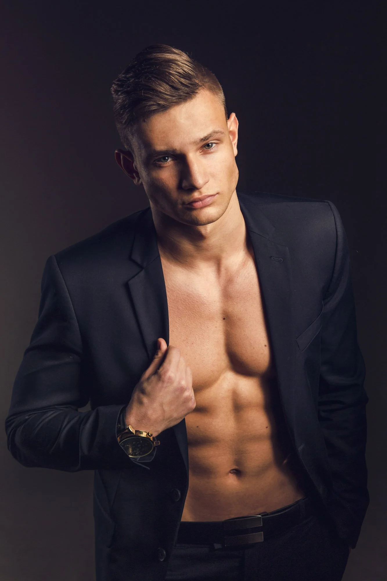 Portrait, Fitness & Commercial Photographer Zurich. 
A shirtless young man with short hair wearing a black blazer, a gold watch, and black pants, posing against a dark background.