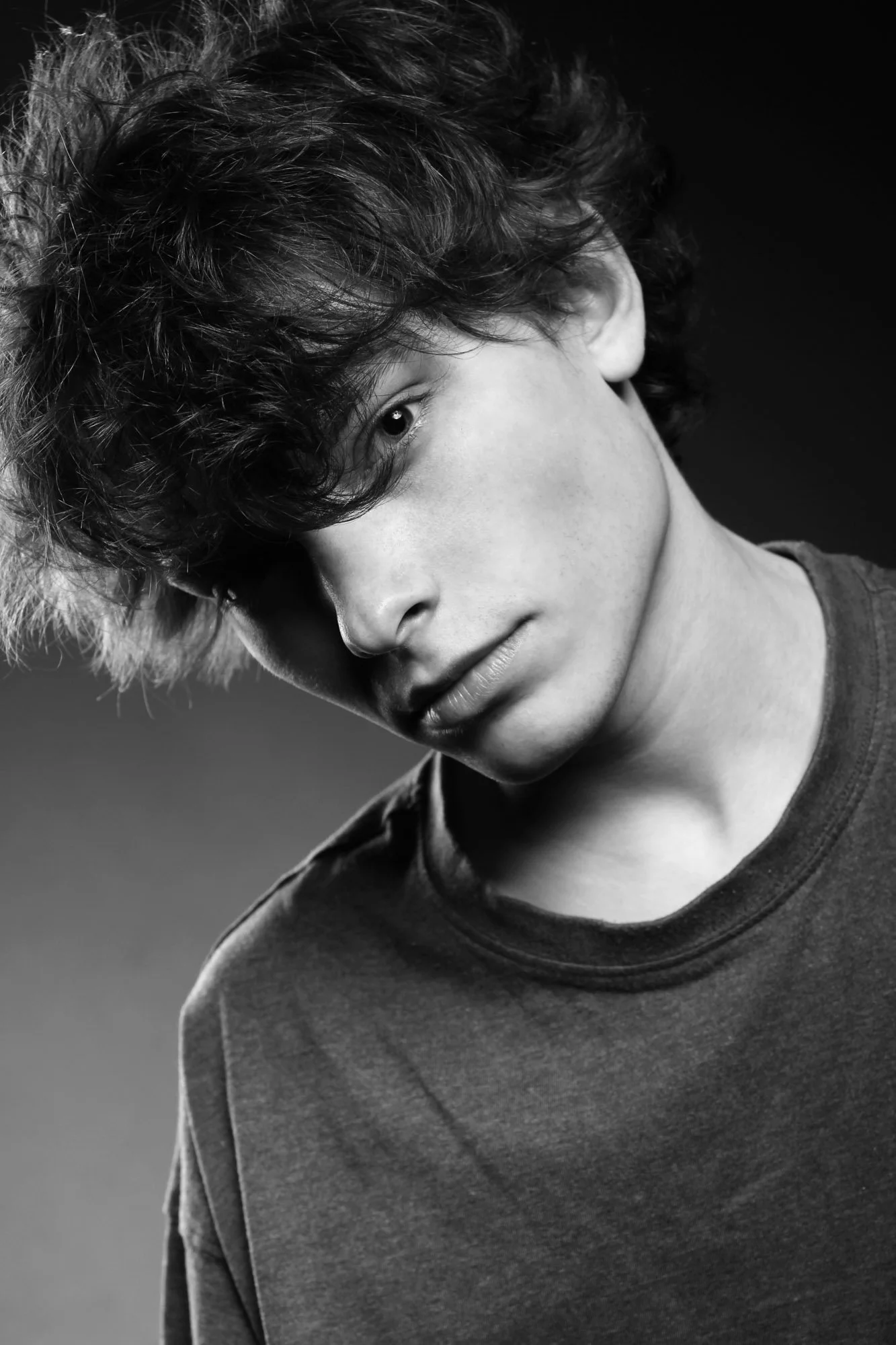 Portrait, Fitness & Commercial Photographer Zurich. Black and white portrait of a young person with curly hair, wearing a dark t-shirt, looking downward with part of their face obscured by hair and shadow.