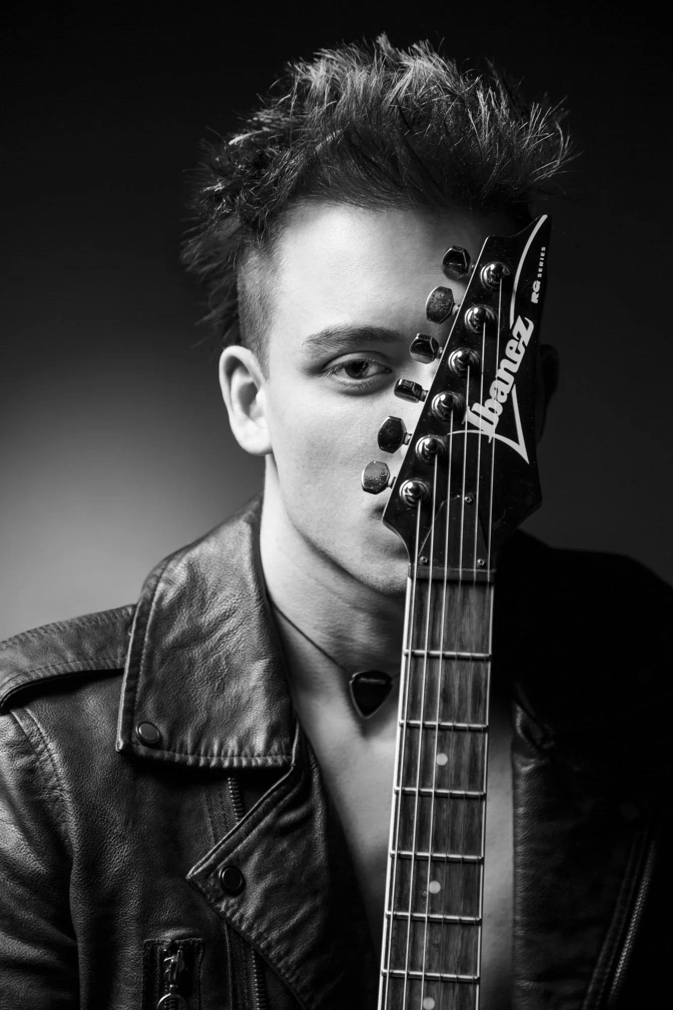Portrait, Fitness & Commercial Photographer Zurich. Black and white photo of a young man with spiky hair looking at the camera, holding an electric guitar partially covering his face, wearing a leather jacket.
