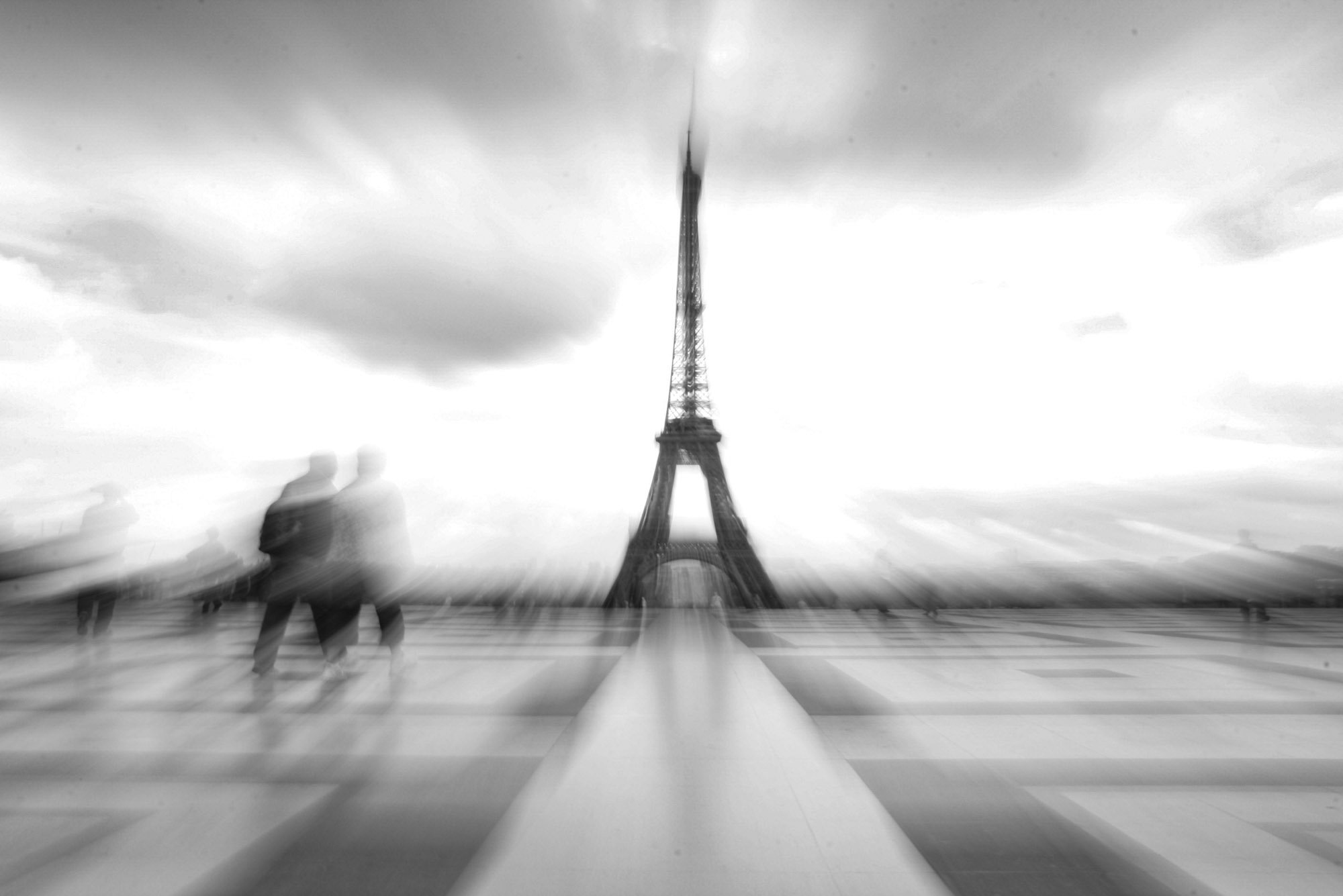Fine art print photography for modern living room wall decor in ZüBlack and white photograph of the Eiffel Tower in Paris, France, with a cloudy sky. Blurred figures of people walking in the foreground. The image appears to have a motion blur effect.