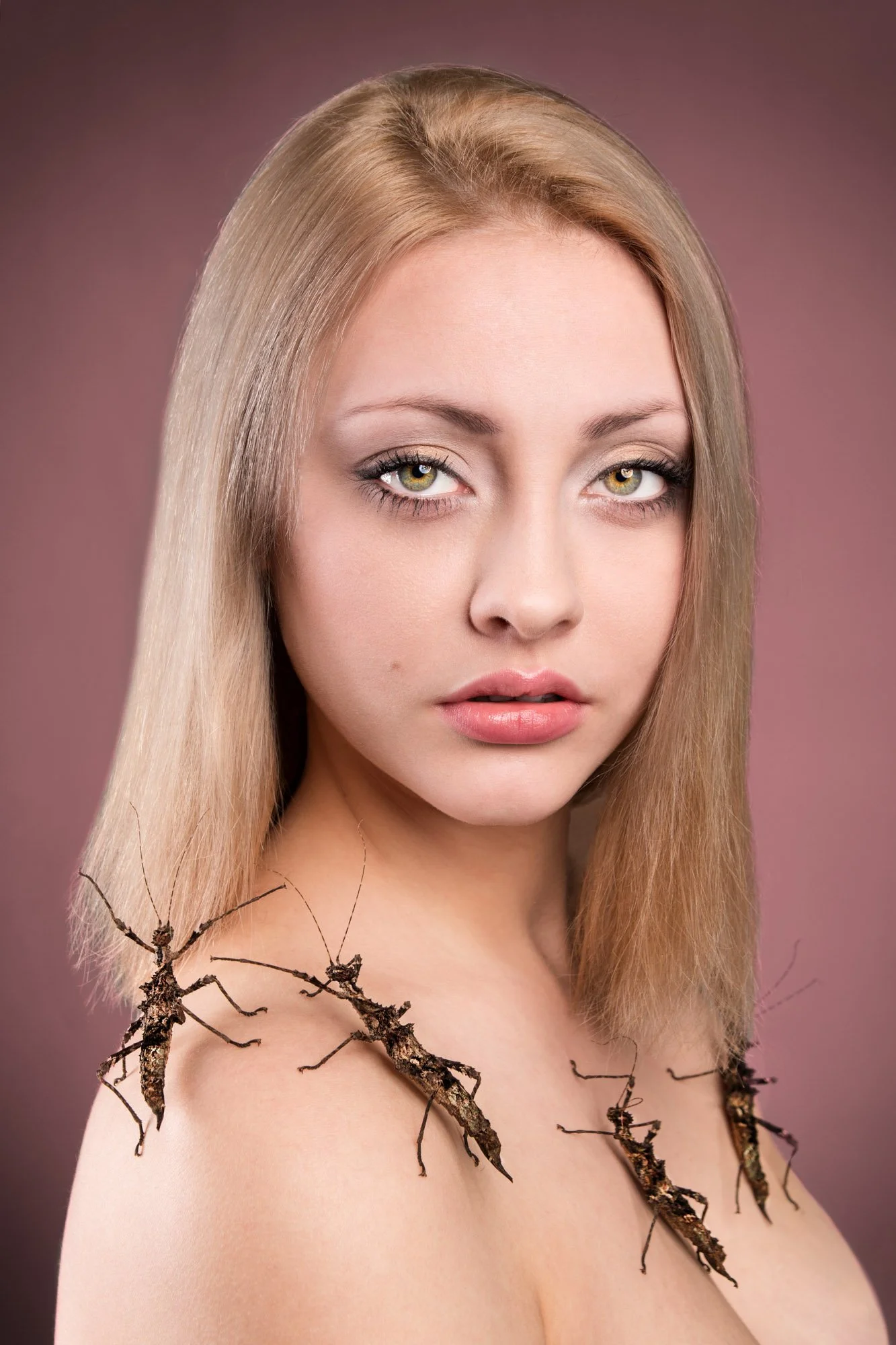 A woman with long blonde hair and light makeup, looking at the camera against a brownish-pink background. Twigs and small branches with dried bark are arranged on her shoulders and upper chest.