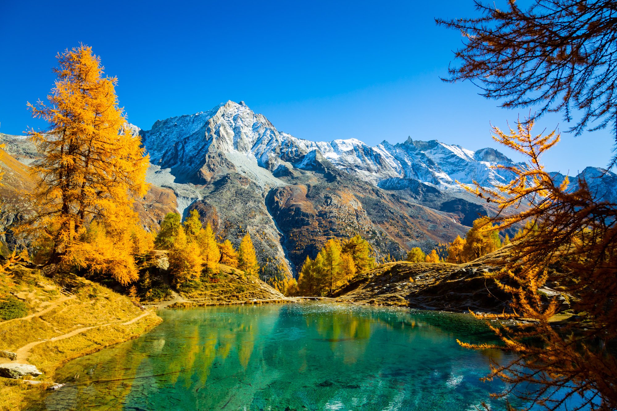 Scenic mountain landscape featuring snow-capped peaks, yellow autumn trees, and a turquoise lake in the foreground under a clear blue sky.