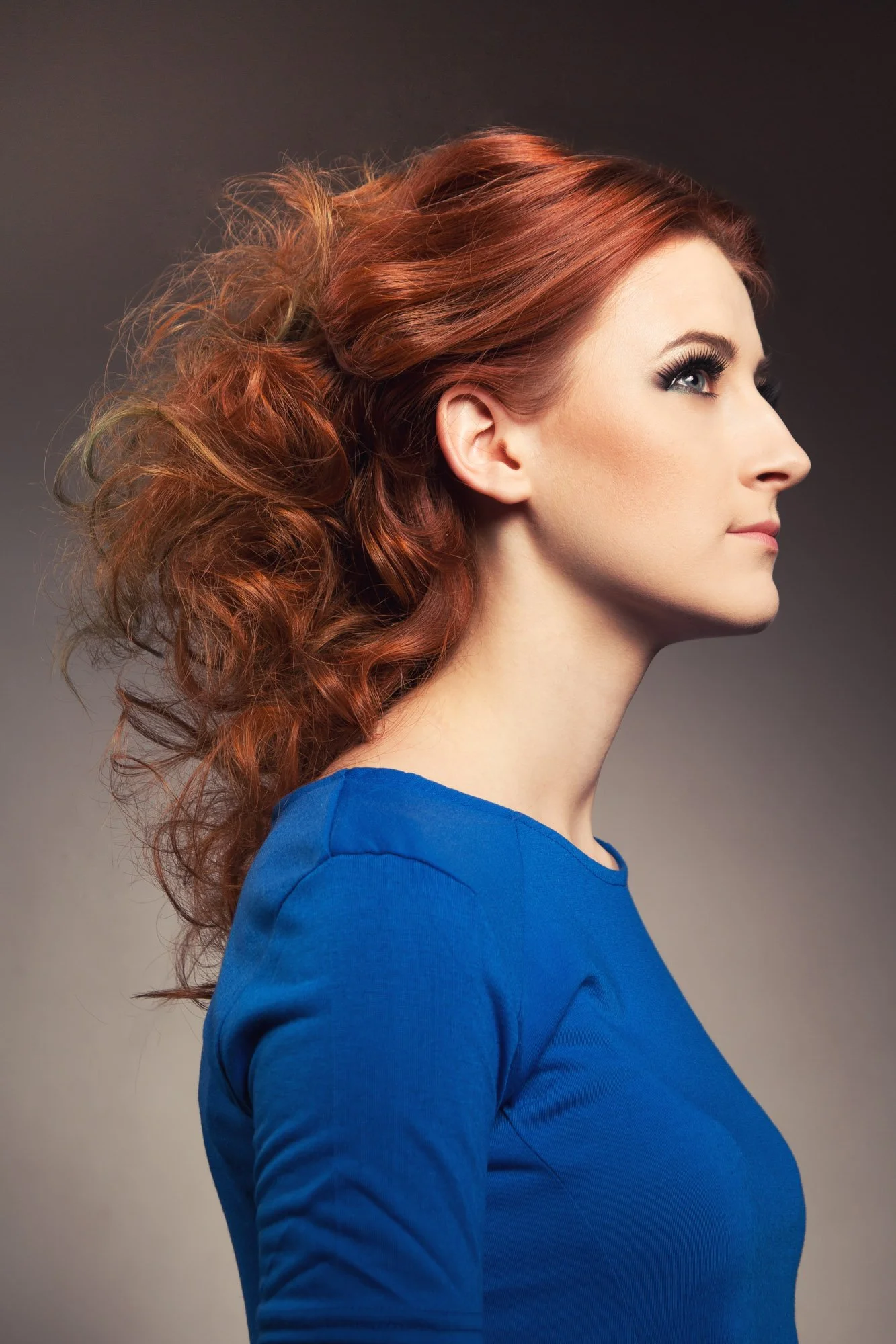 Portrait, Fitness & Commercial Photographer Zurich. Side profile of a woman with curly red hair wearing a blue top against a gradient gray background.