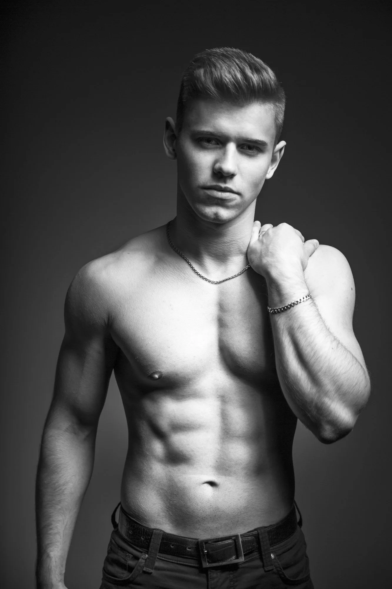 Portrait, Fitness & Commercial Photographer Zurich. Black and white photo of a shirtless young man with short hair, wearing a chain and a bracelet, with his hand on his shoulder, against a plain dark background.