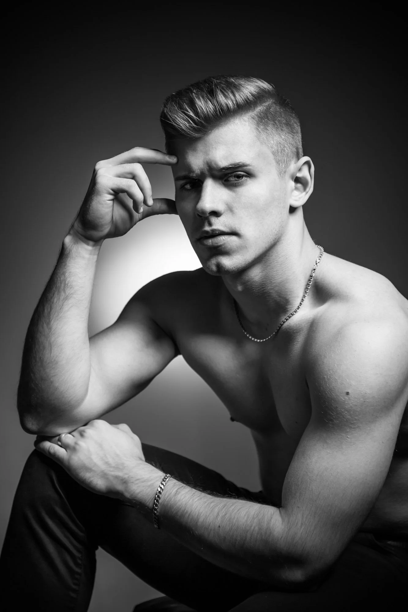 A young man with styled hair, wearing a necklace, seated shirtless against a dark gradient background, looking intently at the camera while resting his head on his hand.