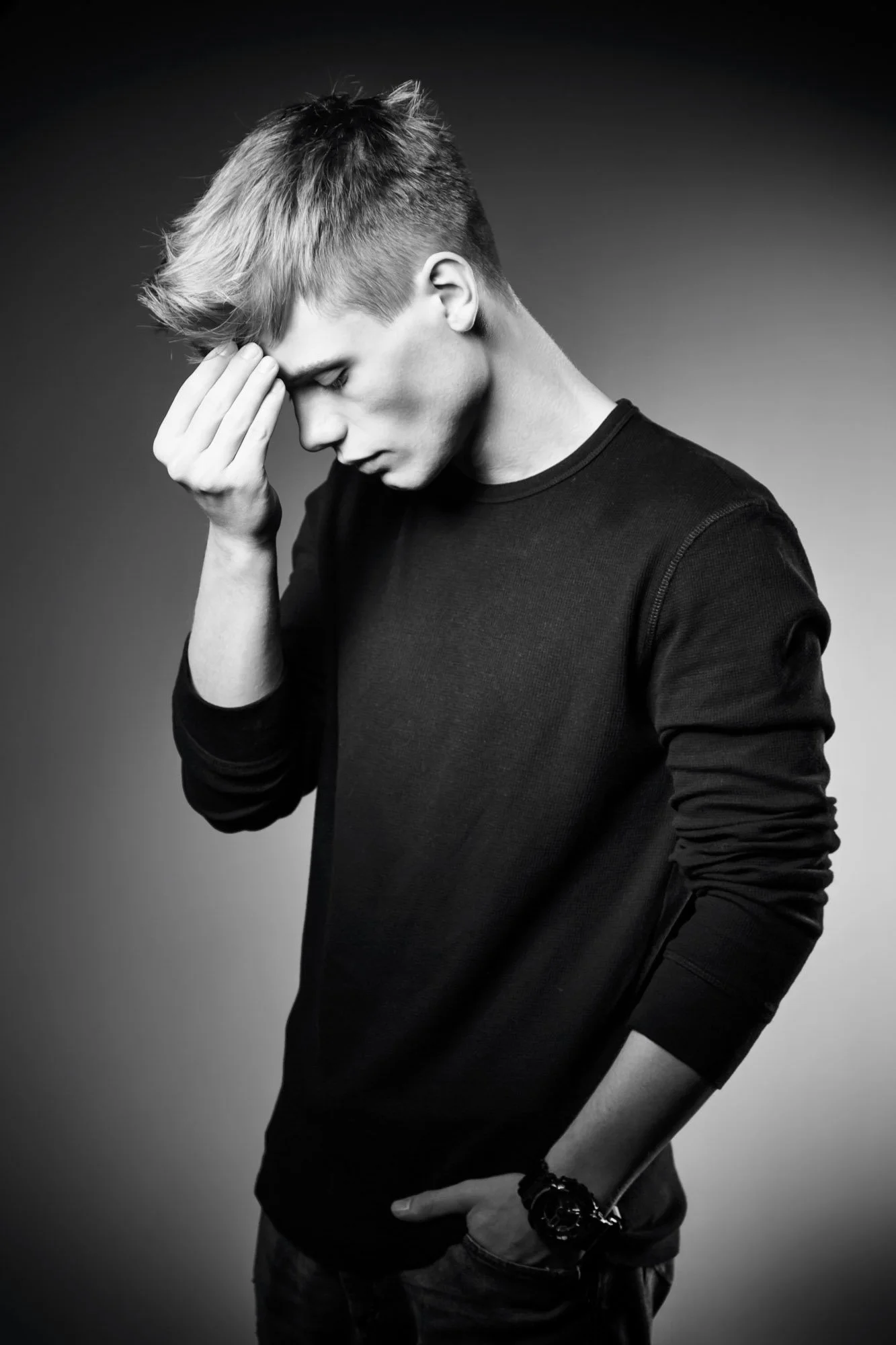 Portrait, Fitness & Commercial PhotographerBlack and white portrait of a young man with a modern haircut, wearing a dark long-sleeve shirt, looking downward with one hand near his forehead and the other resting on his hip, against a plain background.