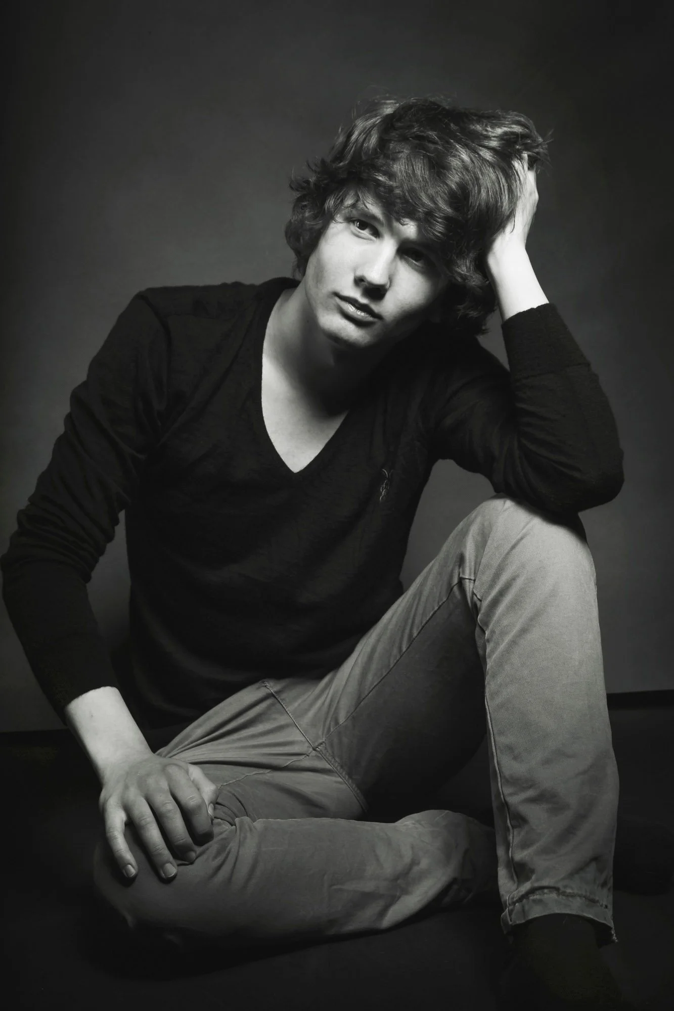 Portrait, Fitness & CommeA black-and-white portrait of a young man with wavy hair, sitting cross-legged on the floor, wearing a long-sleeve shirt and pants, with one hand resting on his knee and the other touching his head, against a dark background.