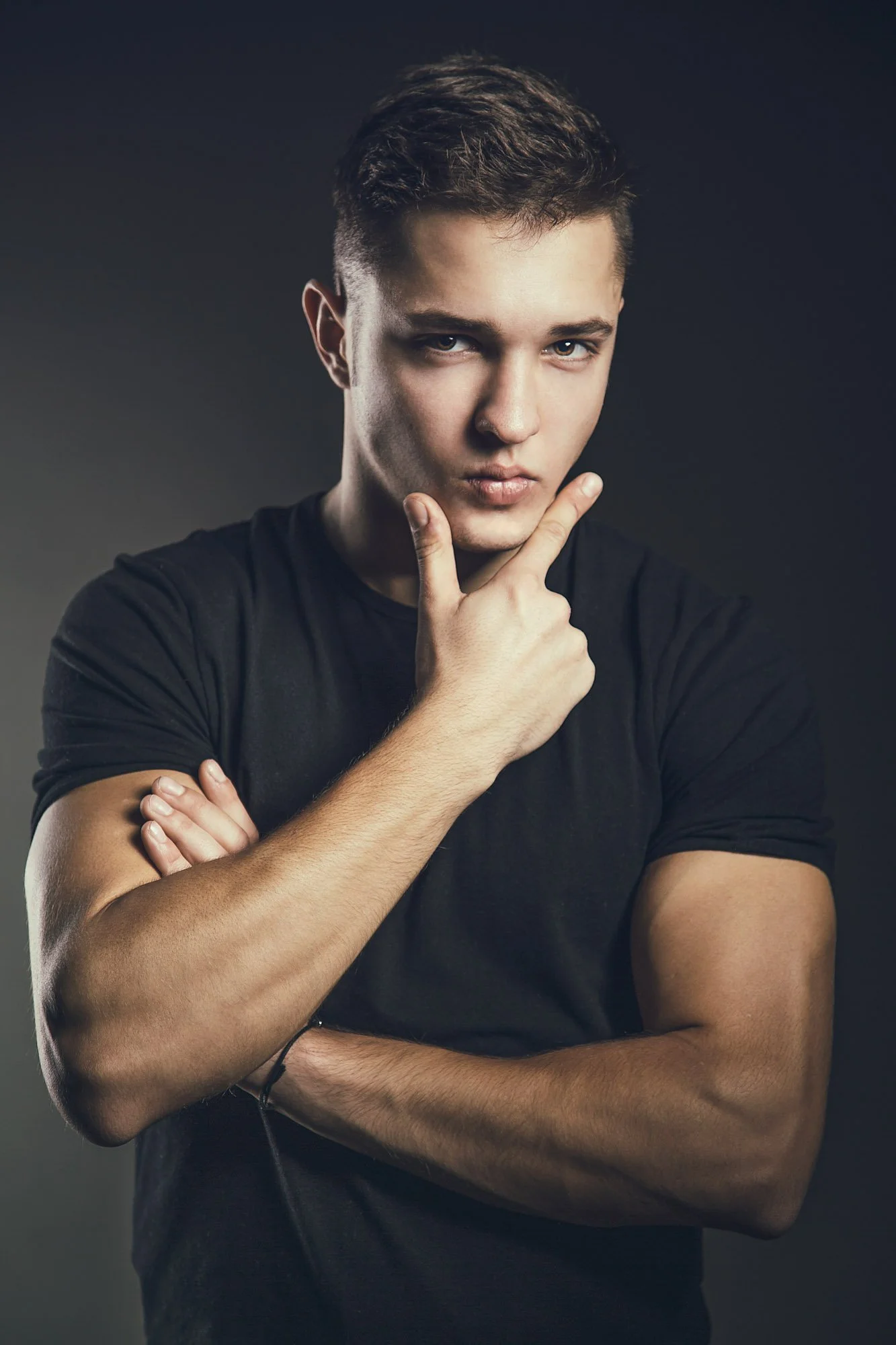 Young man with brown hair and blue eyes, wearing a black t-shirt, looking at the camera with a thoughtful expression, standing against a dark background.