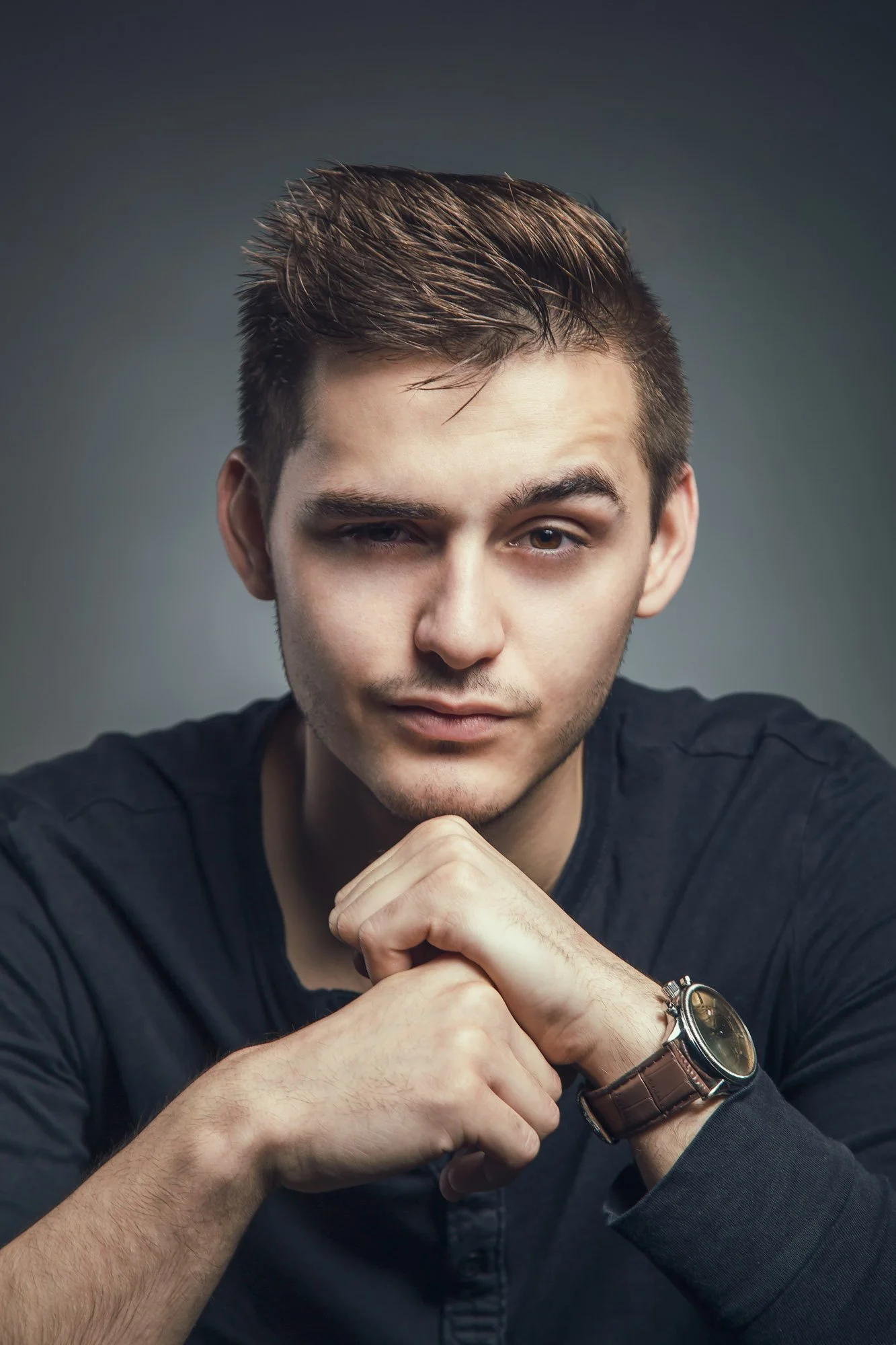 Portrait, Fitness & Commercial Photographer Zurich. A young man with styled brown hair winks at the camera, resting his chin on his hand. He's wearing a dark long-sleeve shirt and a wristwatch with a leather strap. The background is plain and dark.