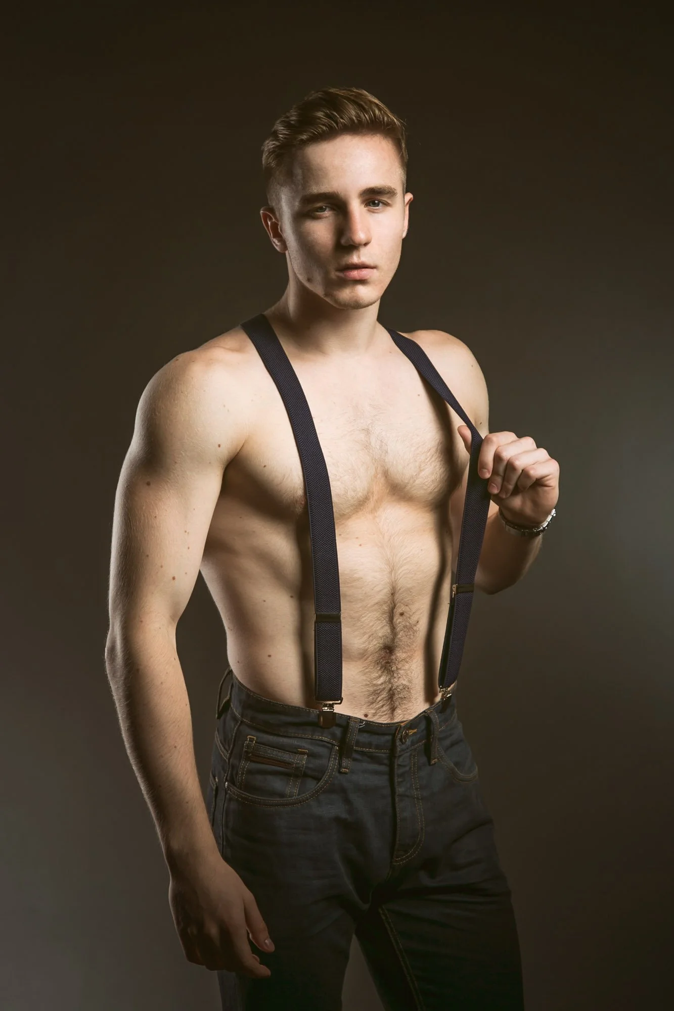 Portrait, Fitness & Commercial Photographer Zurich. A shirtless young man with short hair and a serious expression, holding the straps of suspenders over his shoulder against a dark background.
