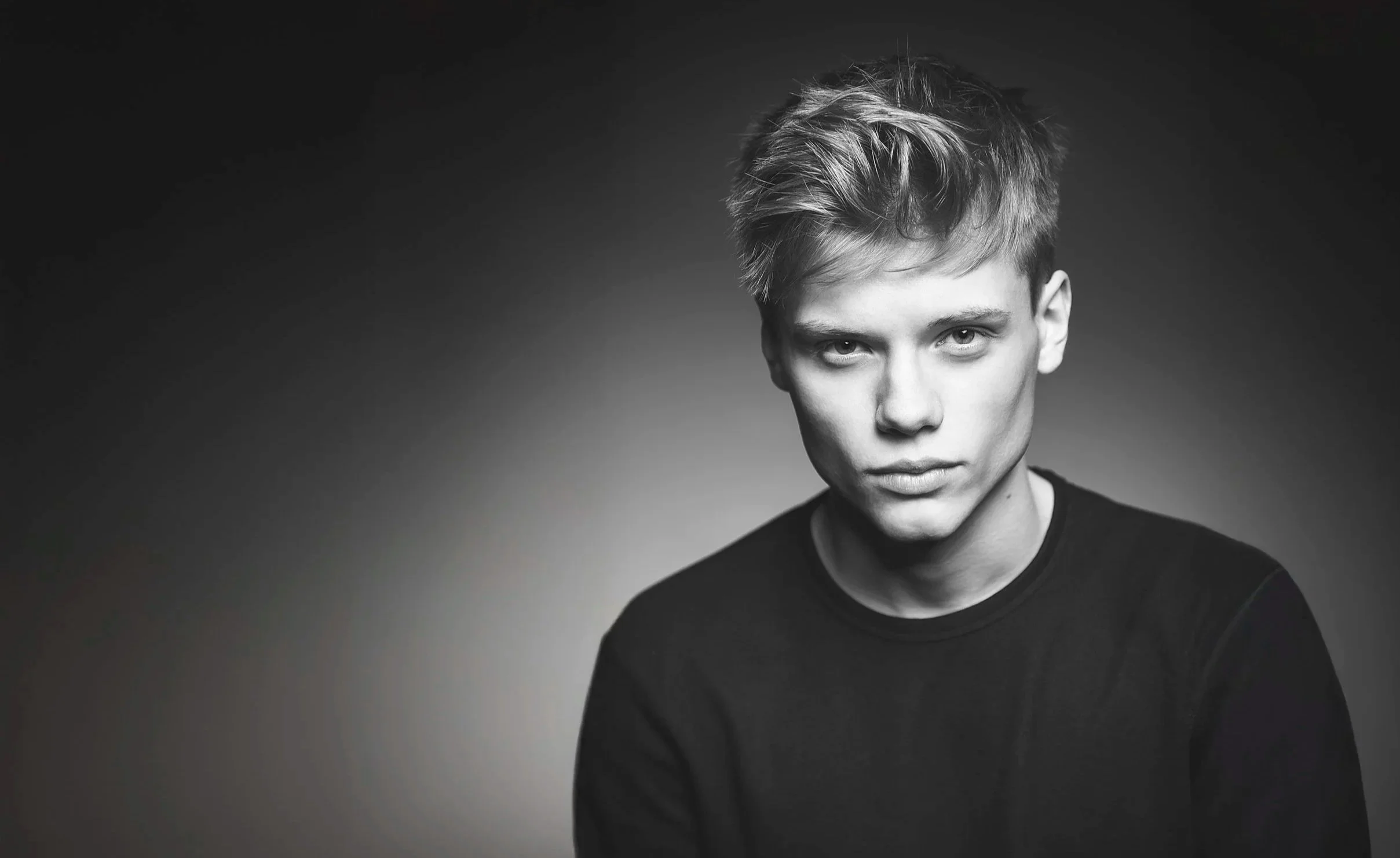 A young man with short, tousled hair and piercing eyes, wearing a black shirt, looking directly at the camera against a dark, gradient background.