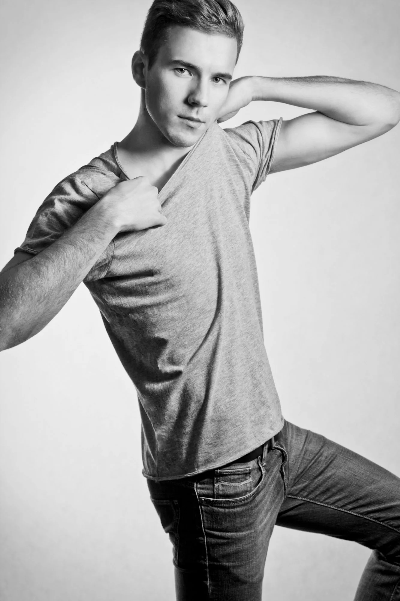 Portrait, Fitness & Commercial Photographer Zurich. A black and white photo of a young man with short hair, wearing a gray t-shirt and jeans, posing with one arm behind his head and the other pulling his shirt.