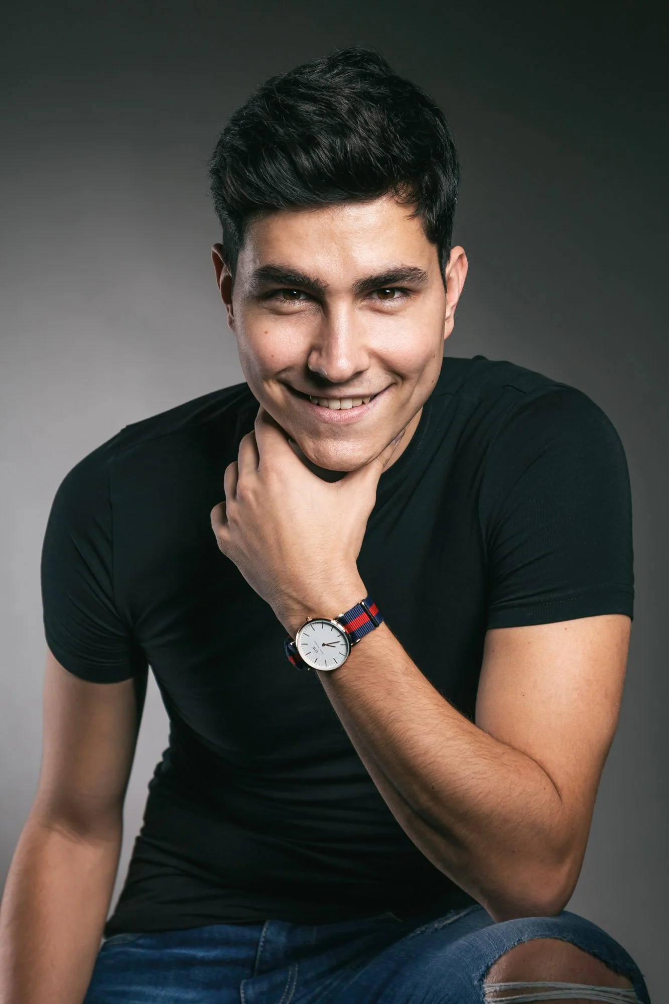 A young man with dark hair and light skin, smiling with his right hand touching his neck, wearing a black t-shirt, a watch, and denim shorts, against a gray background.