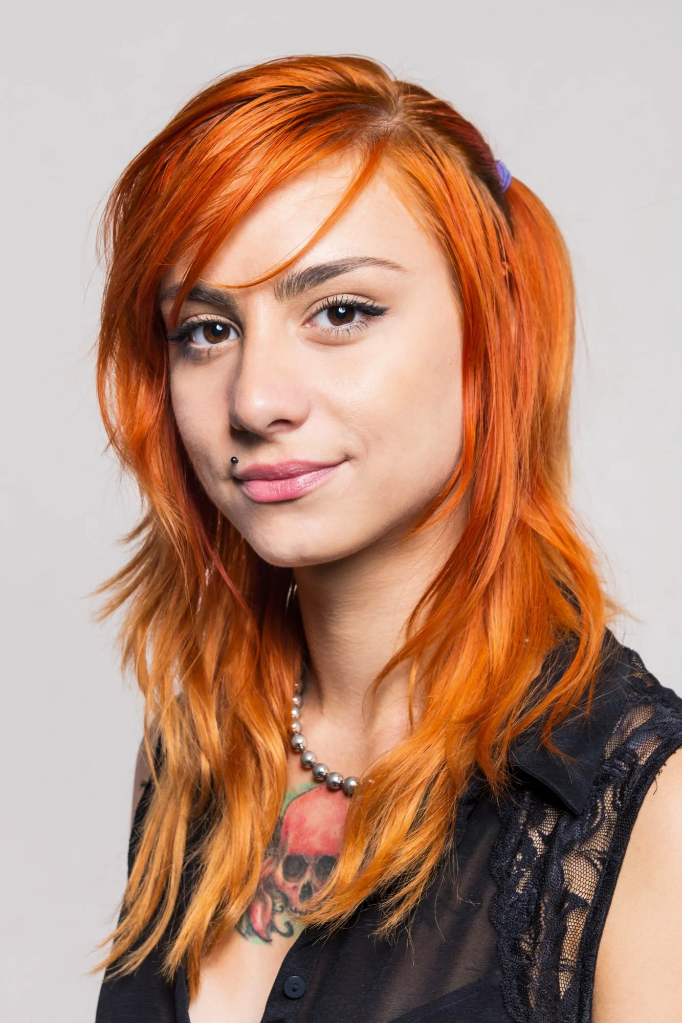 Portrait, Fitness & Commercial Photographer ZurichA young woman with bright orange hair, a small black nose piercing, and a colorful tattoo on her chest, wearing a black lace top and a pearl necklace, smiling slightly against a plain gray background.