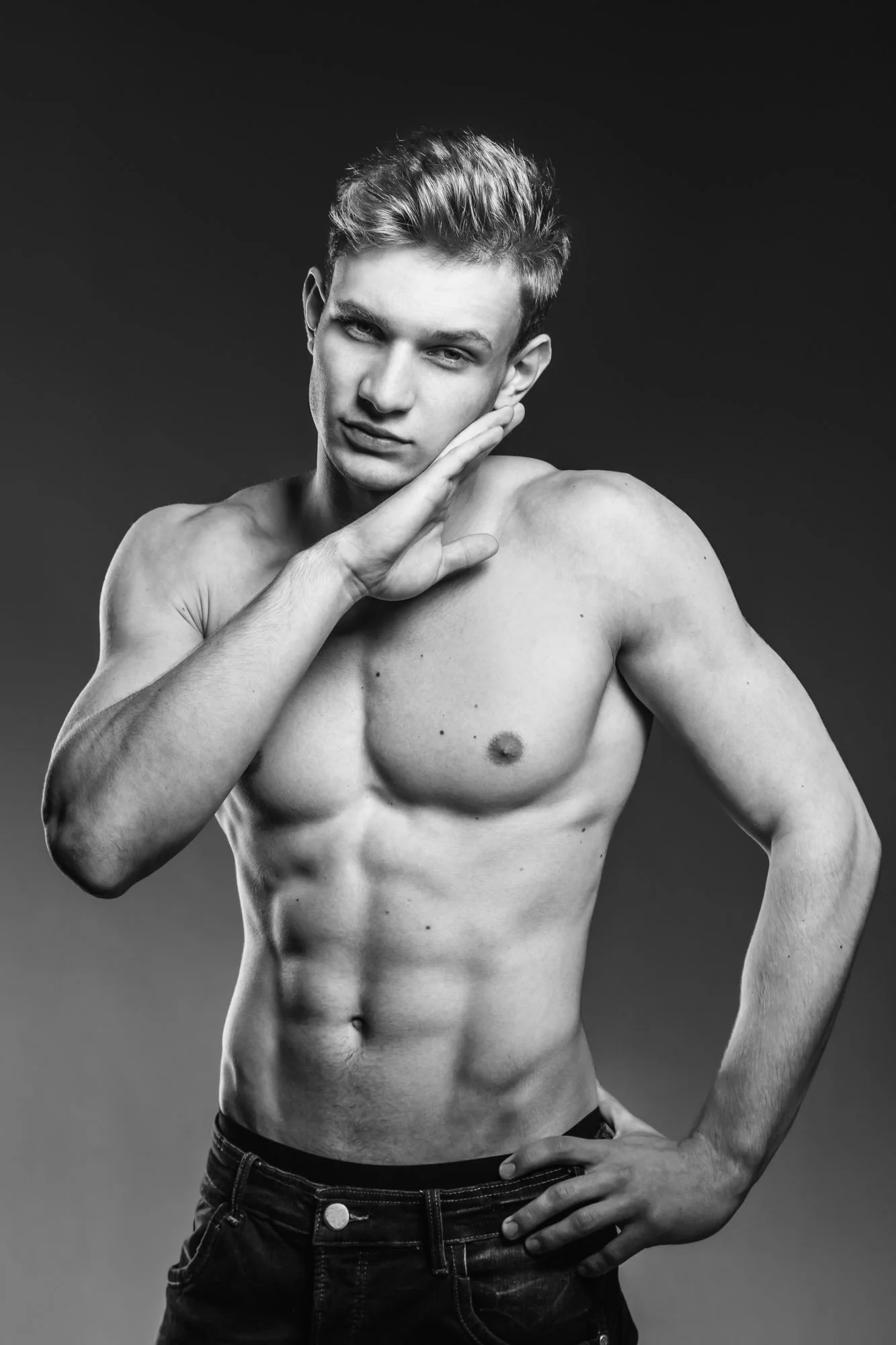 A shirtless young man with styled hair posing against a plain dark background, resting his head on one hand with a confident expression.