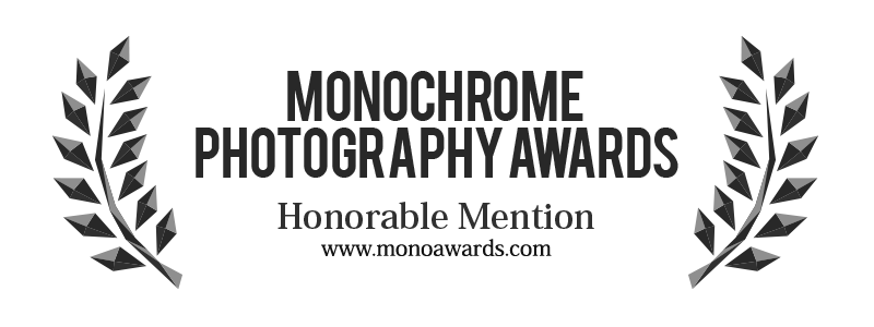 A black award banner for the Monochrome Photography Awards with laurel leaves on both sides, displaying 'Honorable Mention' and the website www.monoawards.com.