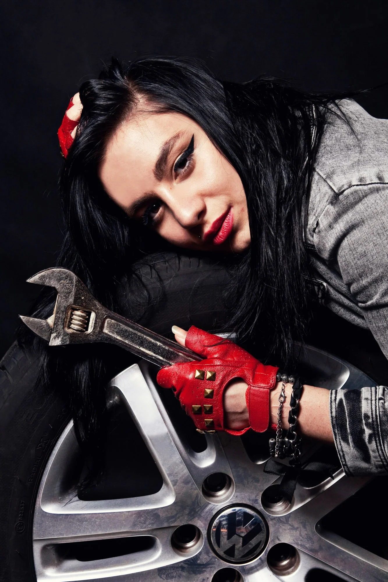 A woman with black hair and red lipstick laying on a tire, holding a wrench. She is wearing red gloves with gold spikes, a gray jacket, and various bracelets. The background is black.