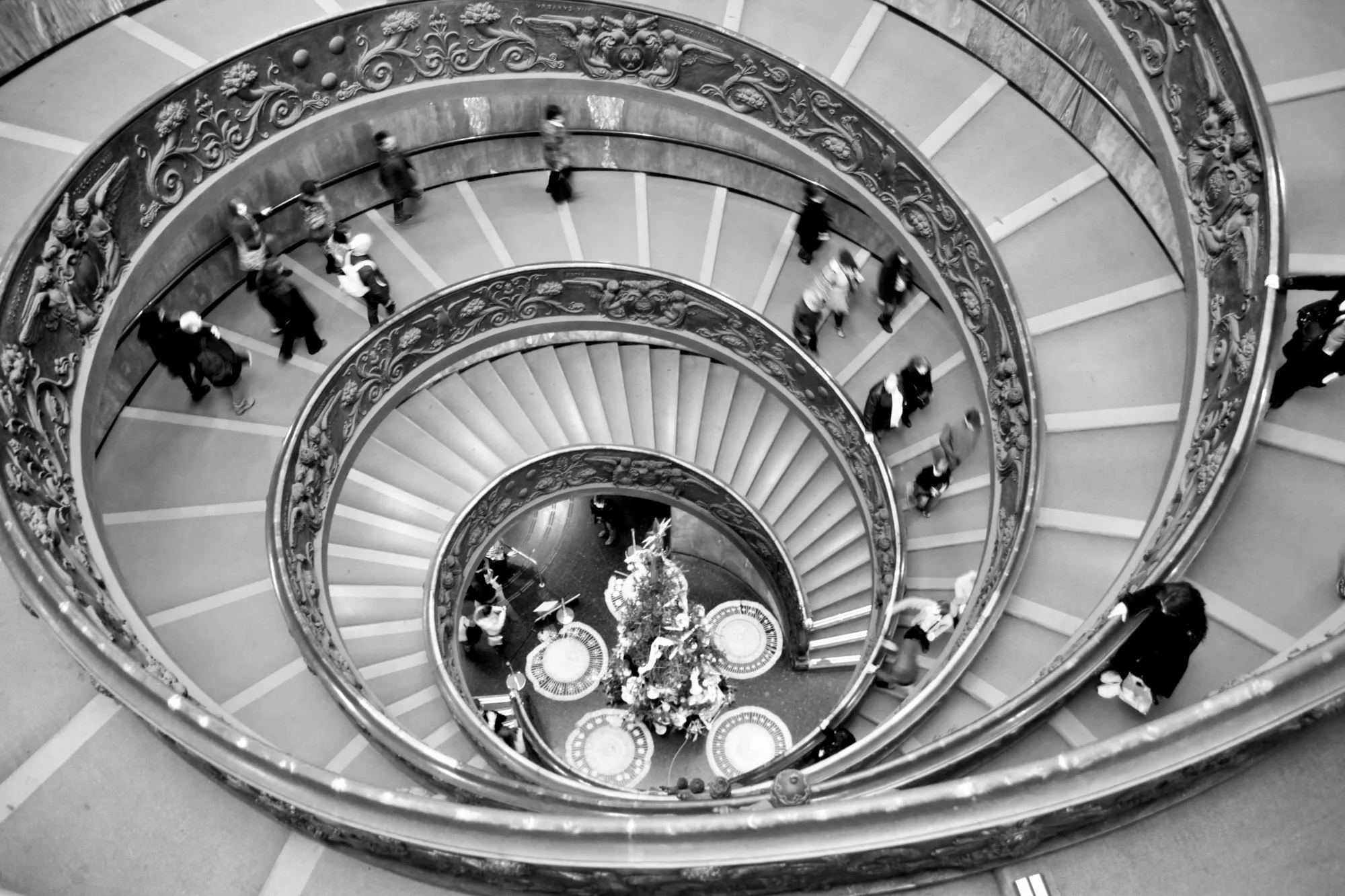 Black and white aerial photograph of a spiral staircase with ornate railing and several people walking on it.