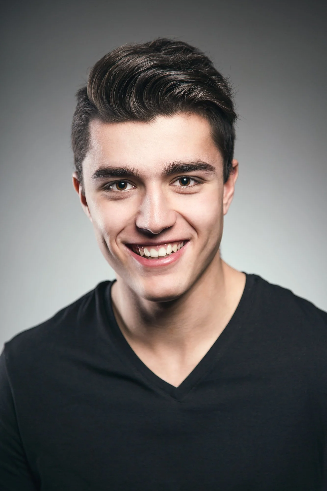Portrait, Fitness & Commercial Photographer Zurich. A young man with dark, styled hair, smiling, wearing a black V-neck shirt, against a plain gray background.