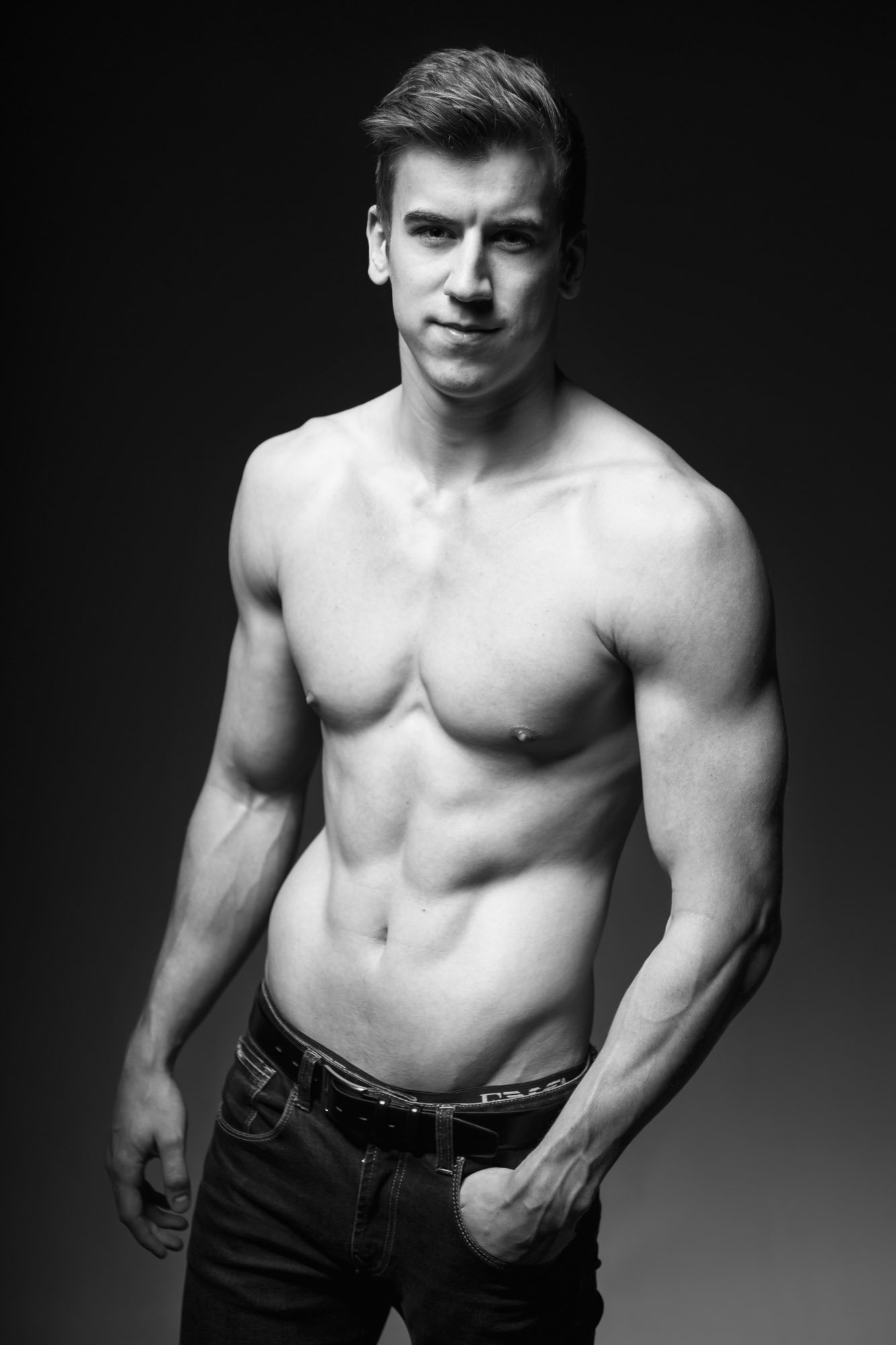 Black and white photo of a shirtless young man with a muscular build, standing against a dark background, with his head slightly tilted and smiling subtly.