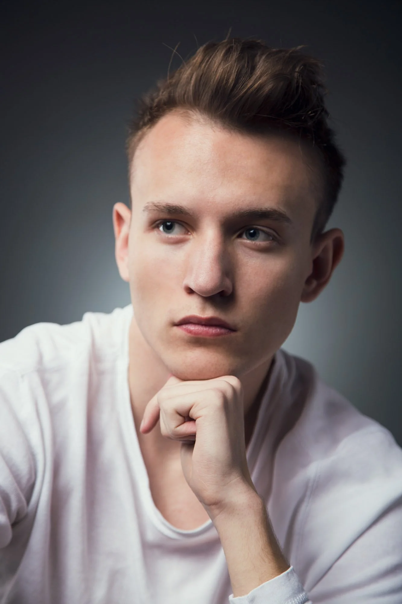 A young man with short brown hair and blue eyes, wearing a white long-sleeve shirt, resting his chin on his hand, looking thoughtfully to the side against a dark background.