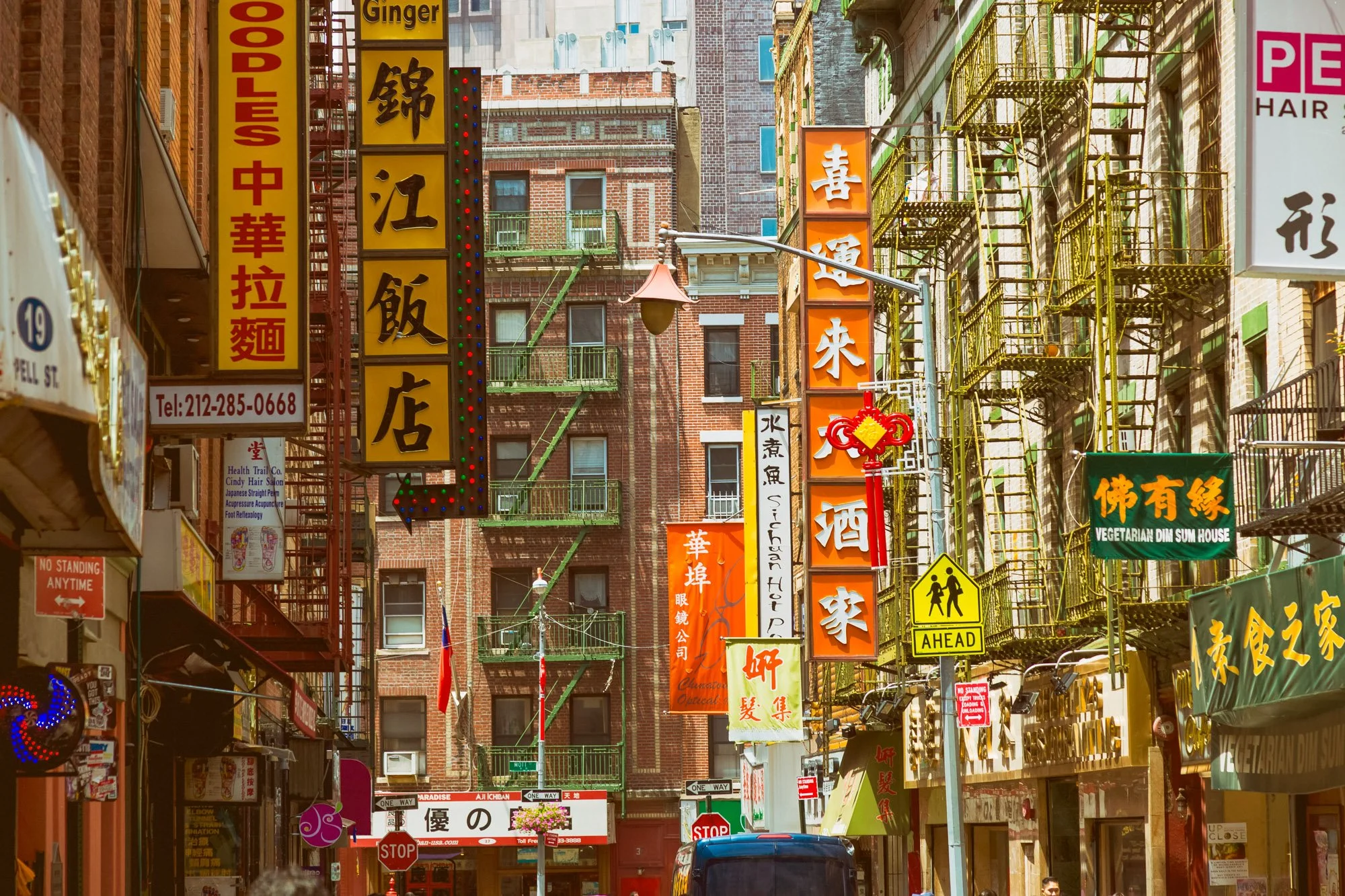 A busy city street in Chinatown with various colorful signs featuring Chinese characters, yellow fire escape ladders on the buildings, and a mix of brick and glass facades. There are street signs and pedestrians visible on the street.