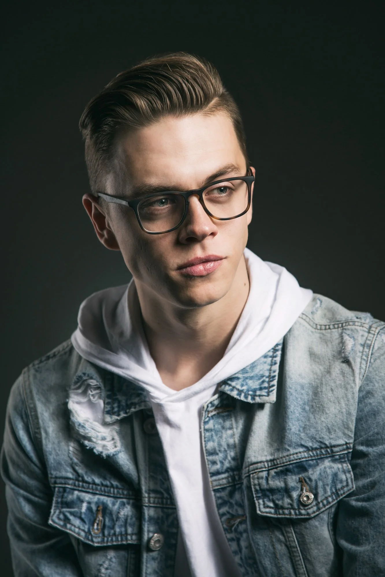 Portrait of a young man with styled hair, glasses, wearing a white hoodie and denim jacket, against a dark background.