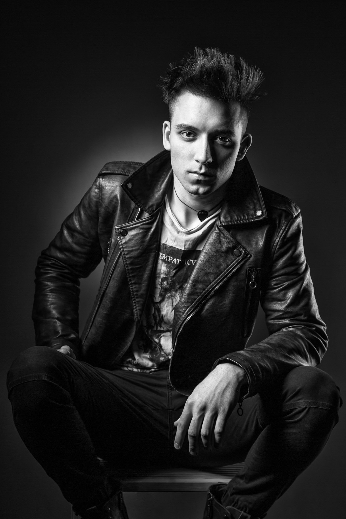 A black and white portrait of a young man with styled hair, wearing a leather jacket and casual clothes, sitting with one hand on his knee against a dark background.