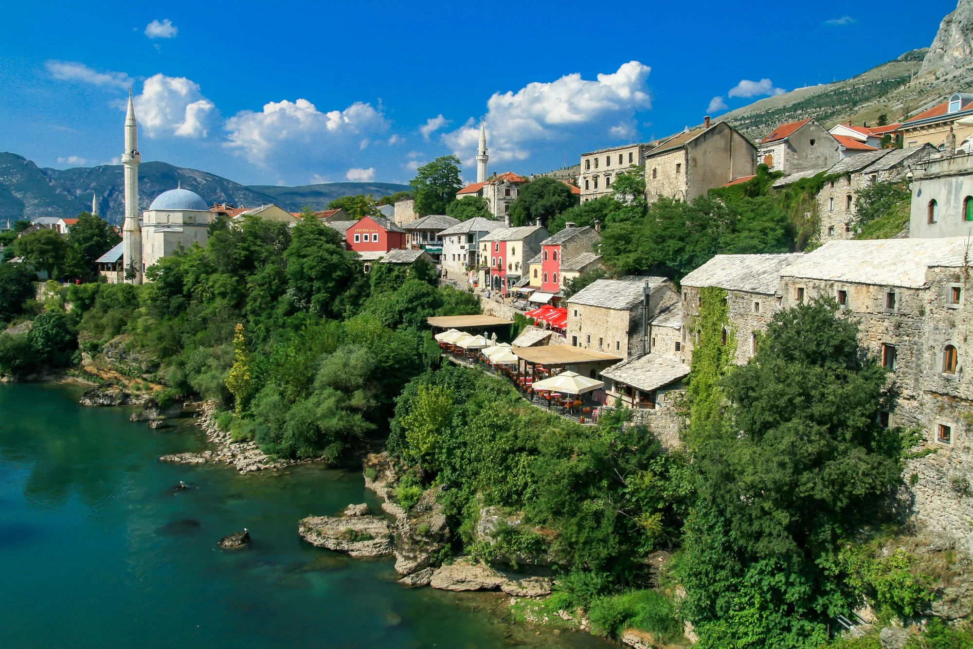 A scenic view of a hillside village with colorful houses, a mosque with minarets, and lush green trees along a winding river under a blue sky with scattered clouds.